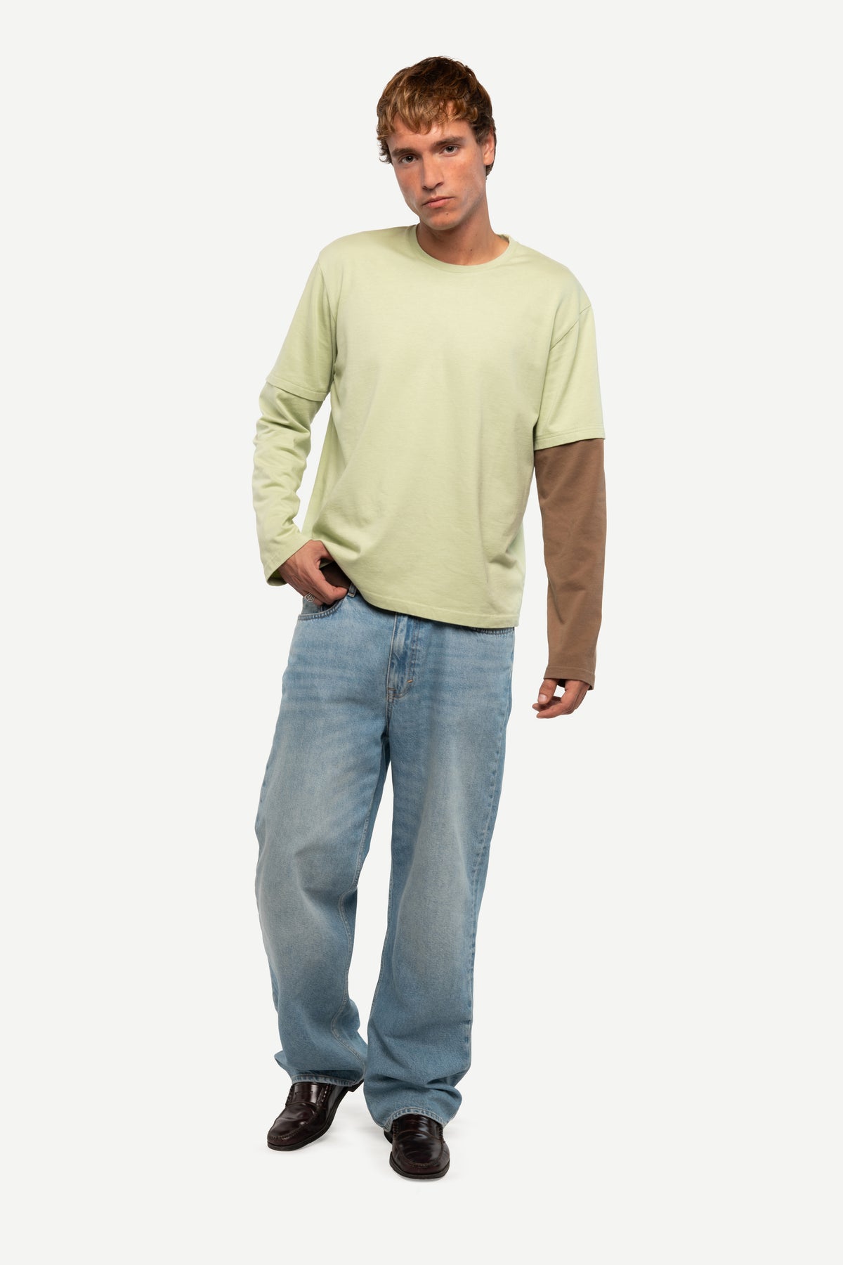 A man modeling a long-sleeve crewneck shirt with a light sage green body and contrasting brown sleeves, paired with light blue jeans and dark brown shoes, against a plain light gray background.