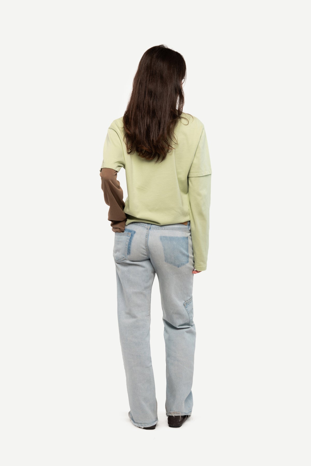 A woman viewed from behind wearing a light sage green long-sleeve top, light blue jeans, and dark brown shoes, set against a plain white studio background.
