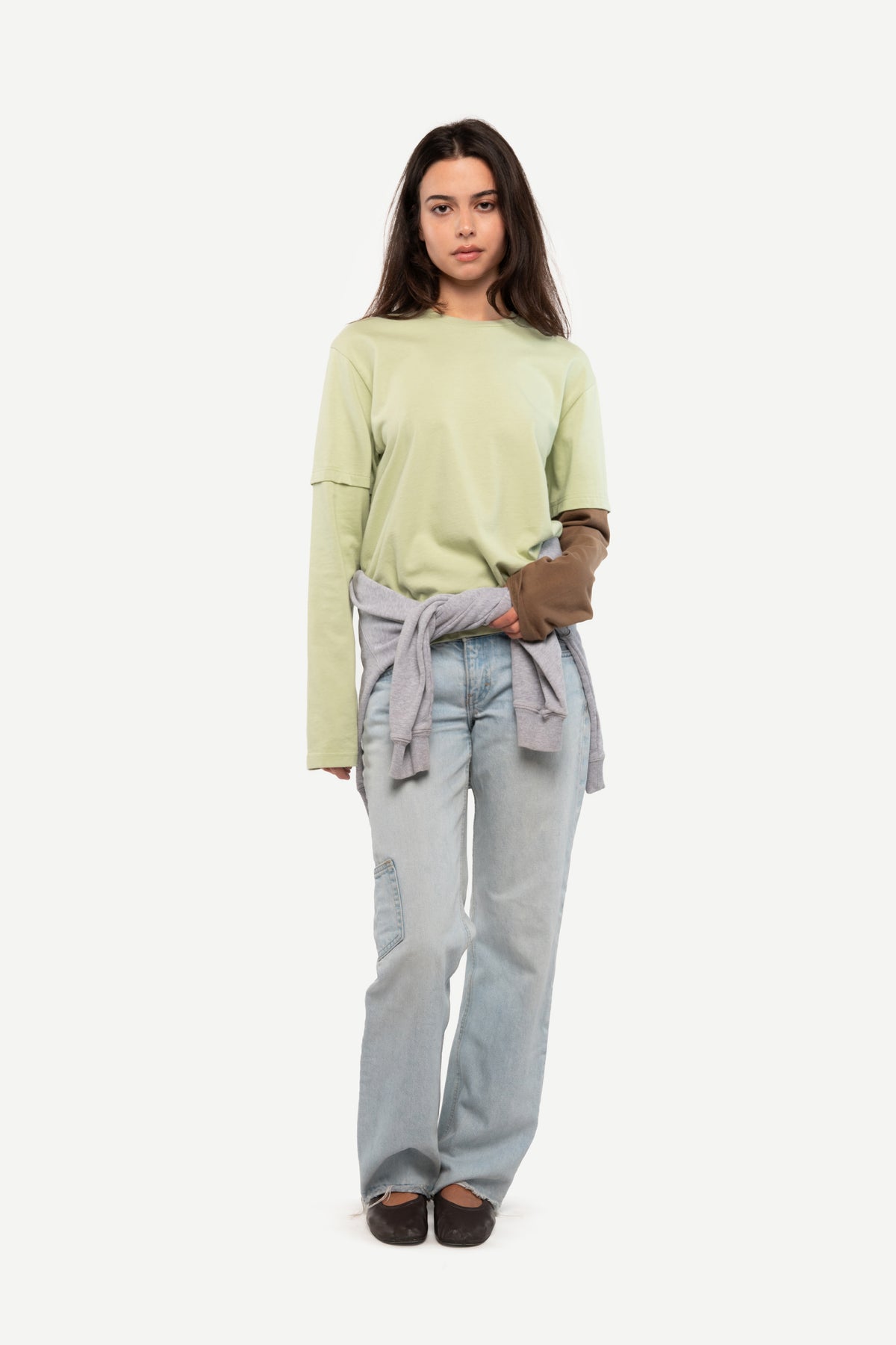 A woman modeling a light sage green long-sleeve top, light-wash blue jeans, and a gray sweater tied around her waist, standing against a plain light gray background.