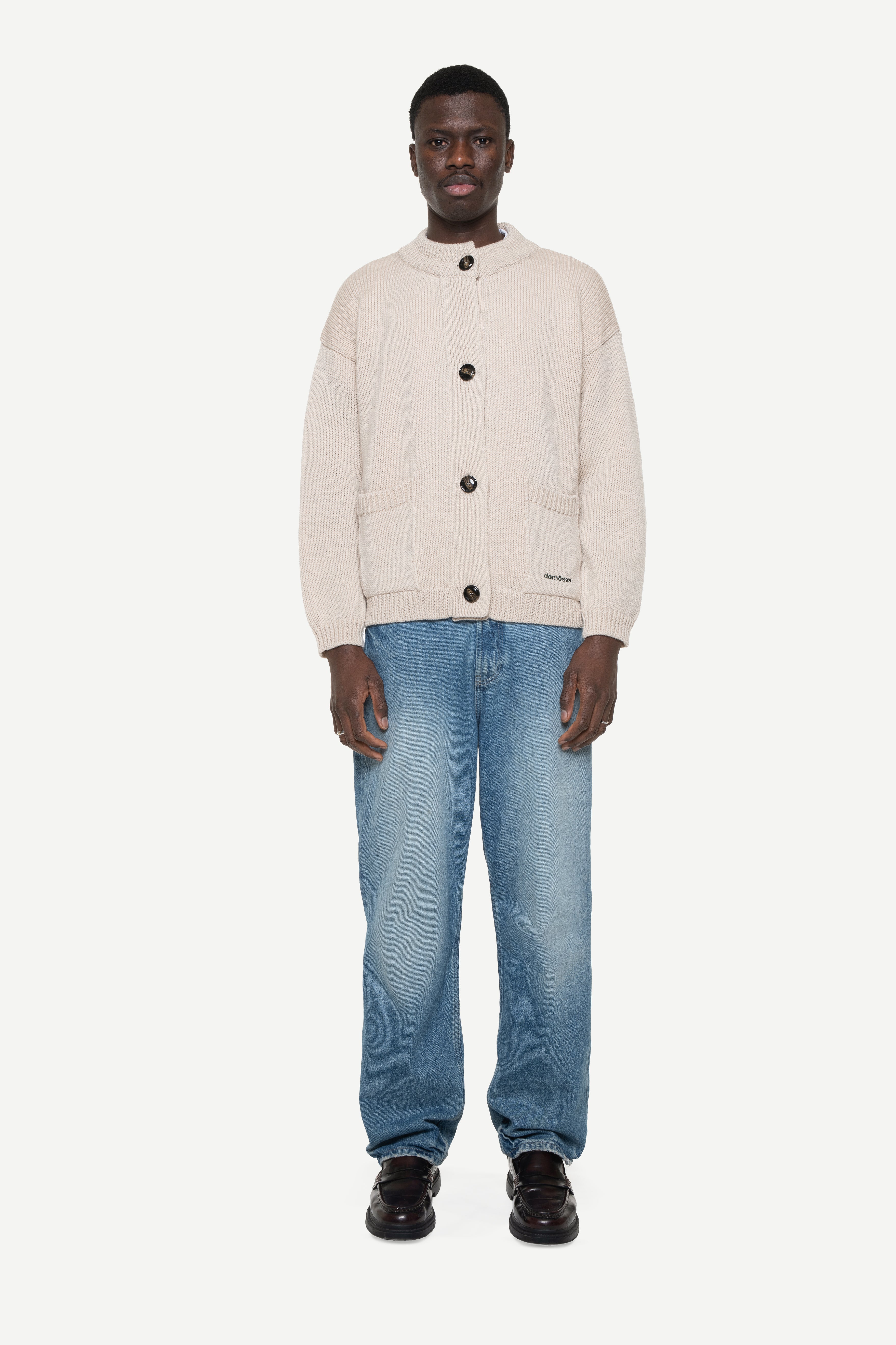 man wearing a beige buttoned cardigan with two front pockets and jeans on a white background
