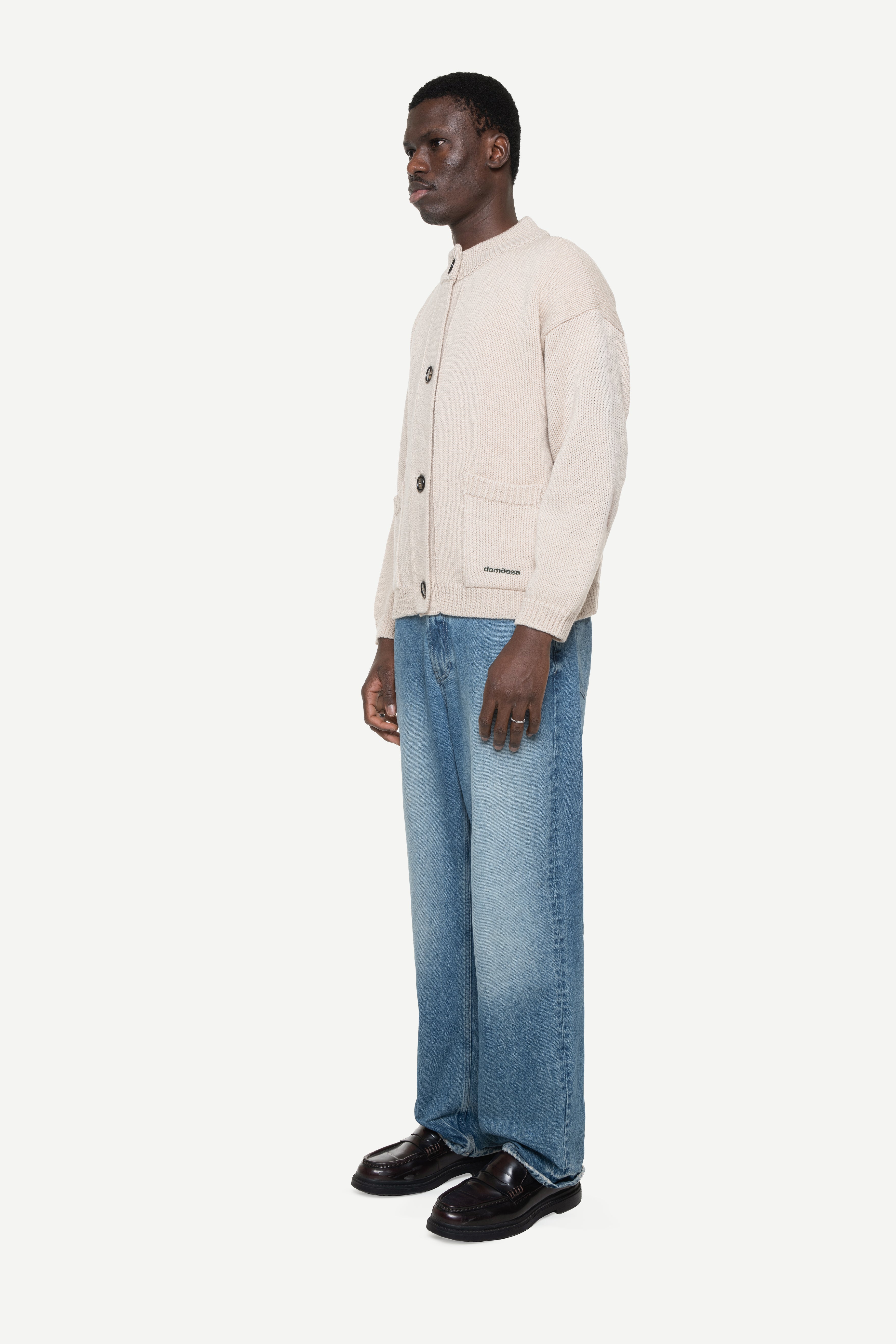 side view of a man wearing a beige buttoned cardigan with two front pockets and jeans on a white background