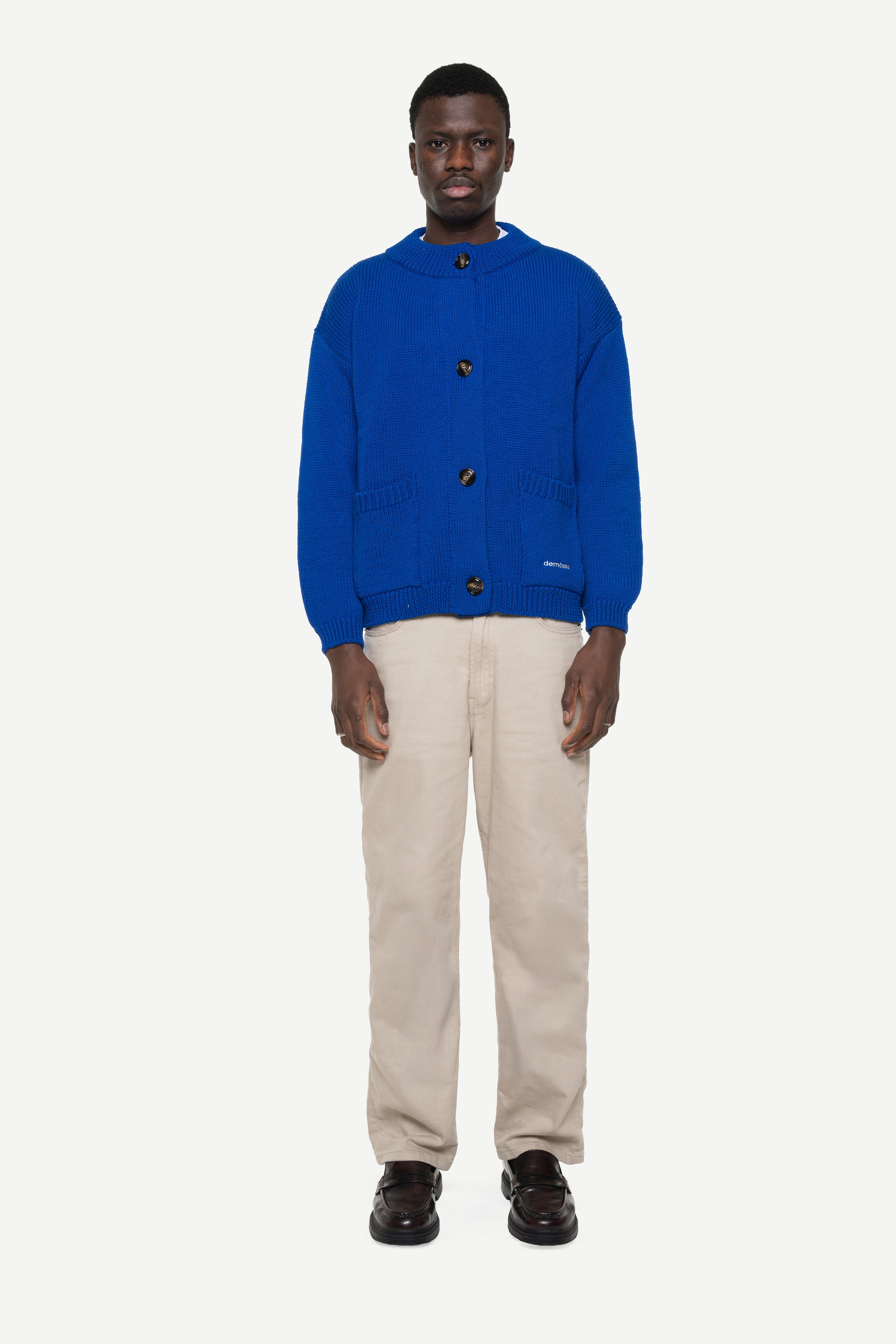 Man wearing a blue knit cardigan with two front pockets and beige pants on a white background