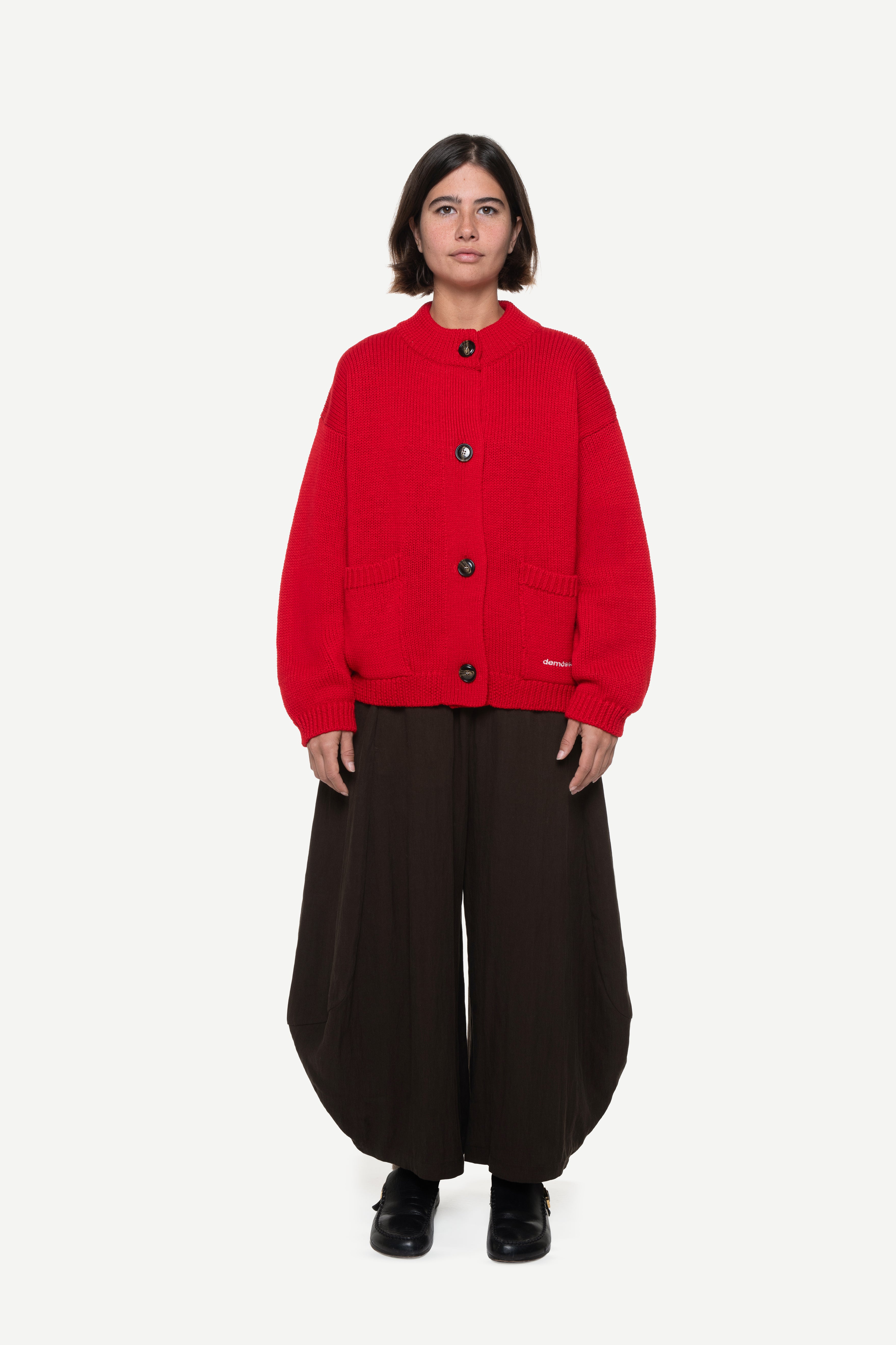 Woman wearing a red buttoned cardigan with two front pockets and brown pants on a white background