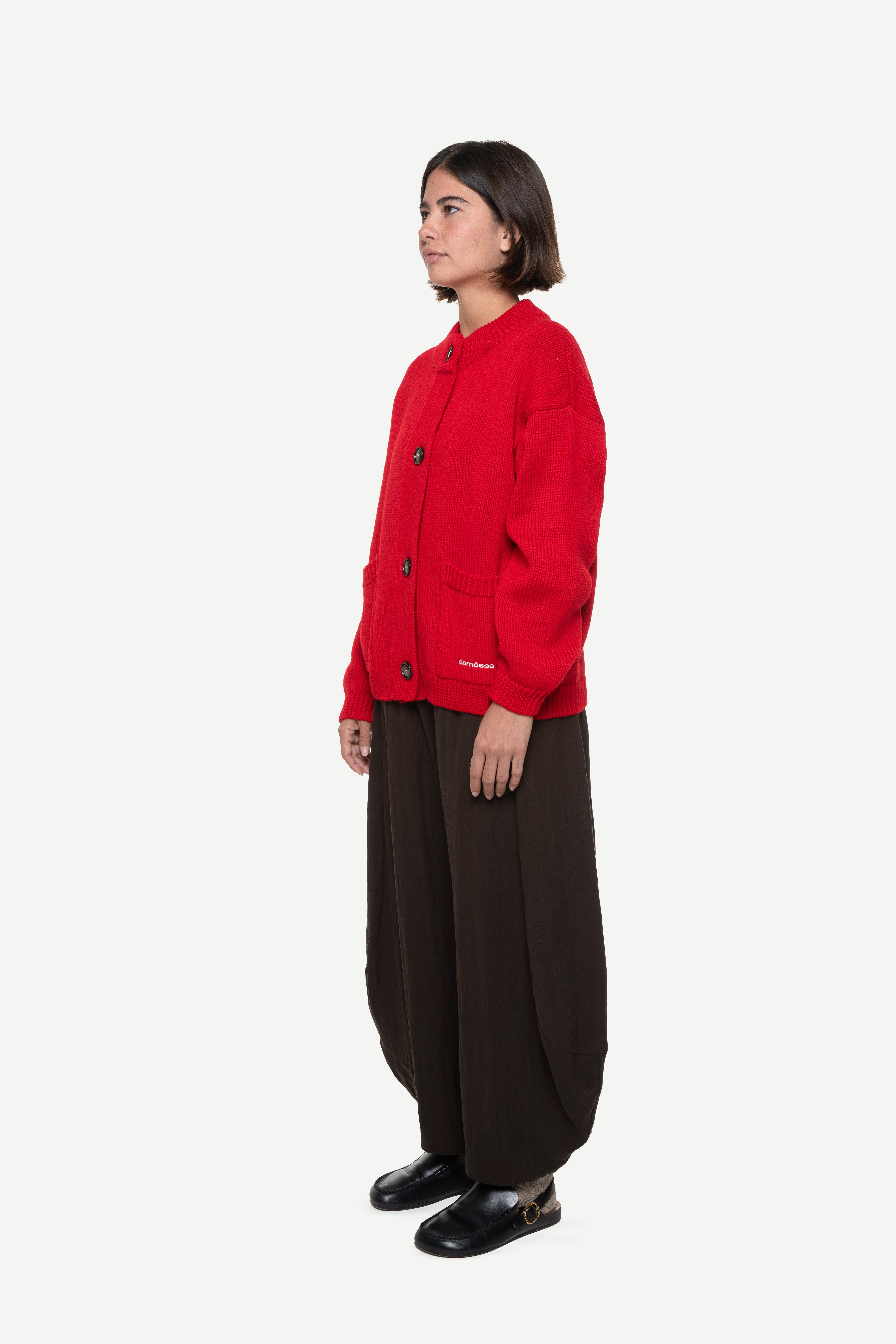 Side view of a woman wearing a red buttoned cardigan with two front pockets and brown pants on a white background