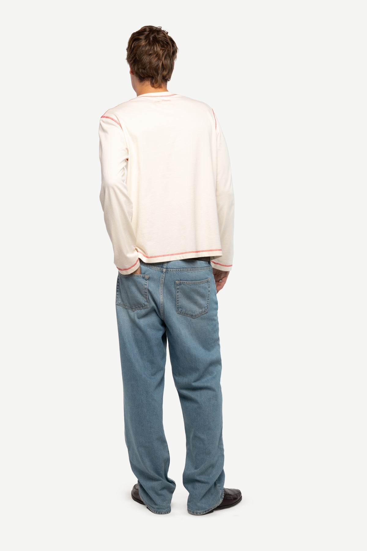 A man viewed from behind wearing a cream-colored long-sleeve bomber jacket with thin red stripes on the shoulders and hem, paired with blue jeans and dark shoes, against a light gray studio background.