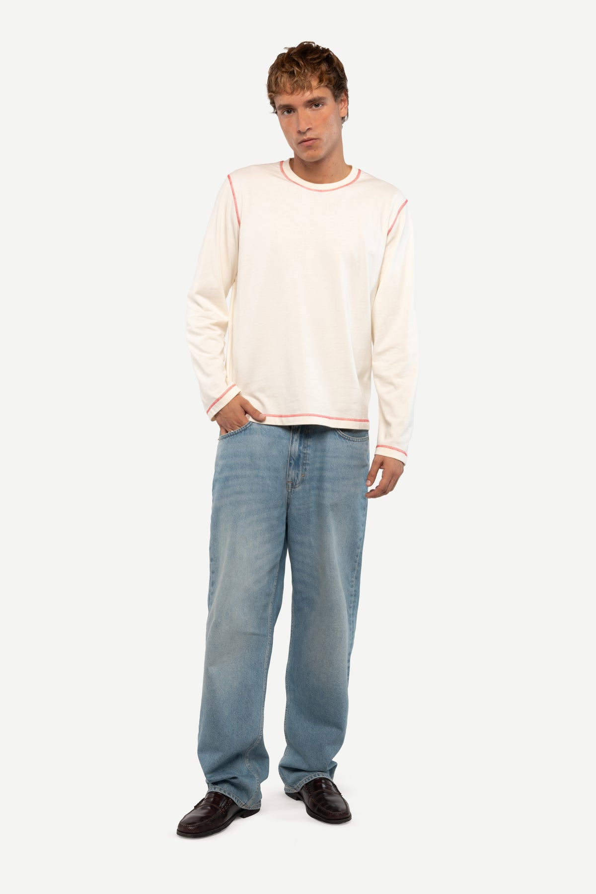 A man modeling a cream long-sleeve knit top with thin orange-red piping details, paired with light blue jeans and dark brown shoes, against a solid off-white background.