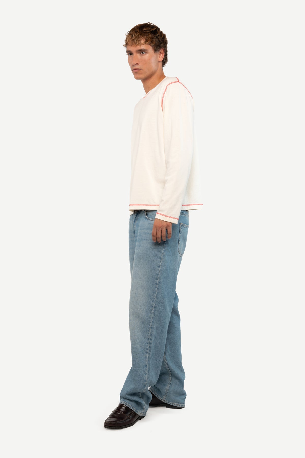 A man wearing a cream long-sleeve knit sweater with thin red stripes on the shoulders and cuffs, paired with light blue jeans and black shoes, standing against a plain light gray studio background.