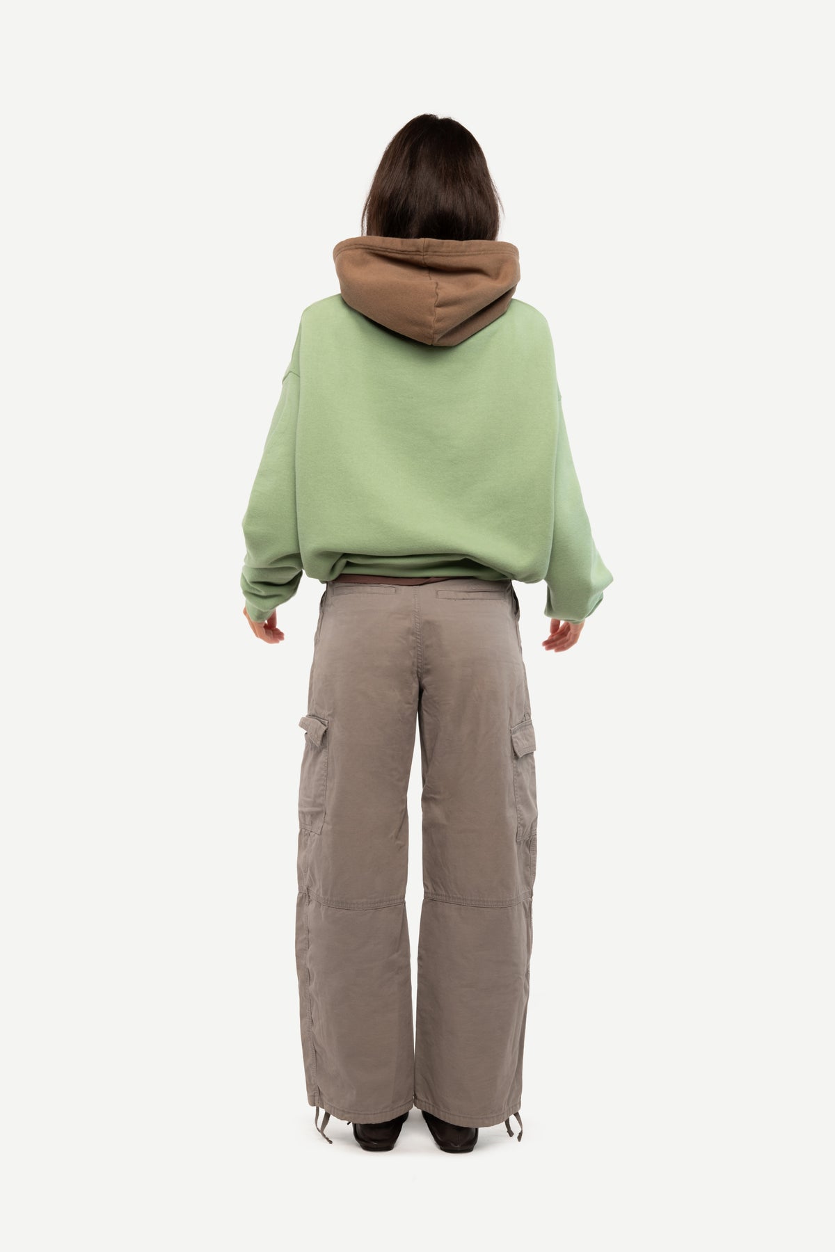 A model shown from the back wearing a light green oversized hoodie with a brown hood lining and wide-leg taupe cargo pants, set against a plain light gray background.