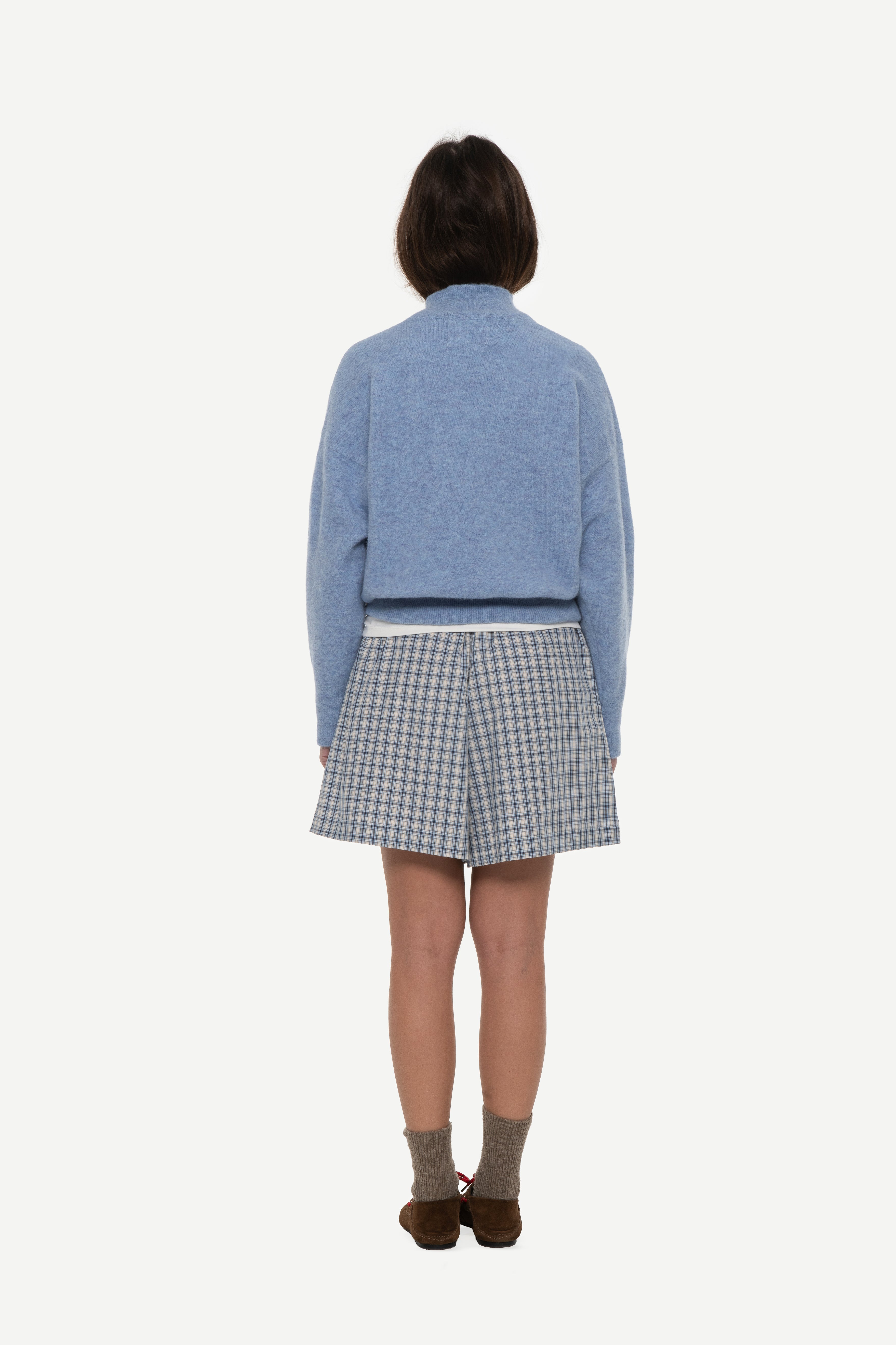 Back view of woman wearing a baby blue mockneck sweater and blue shorts on a white background