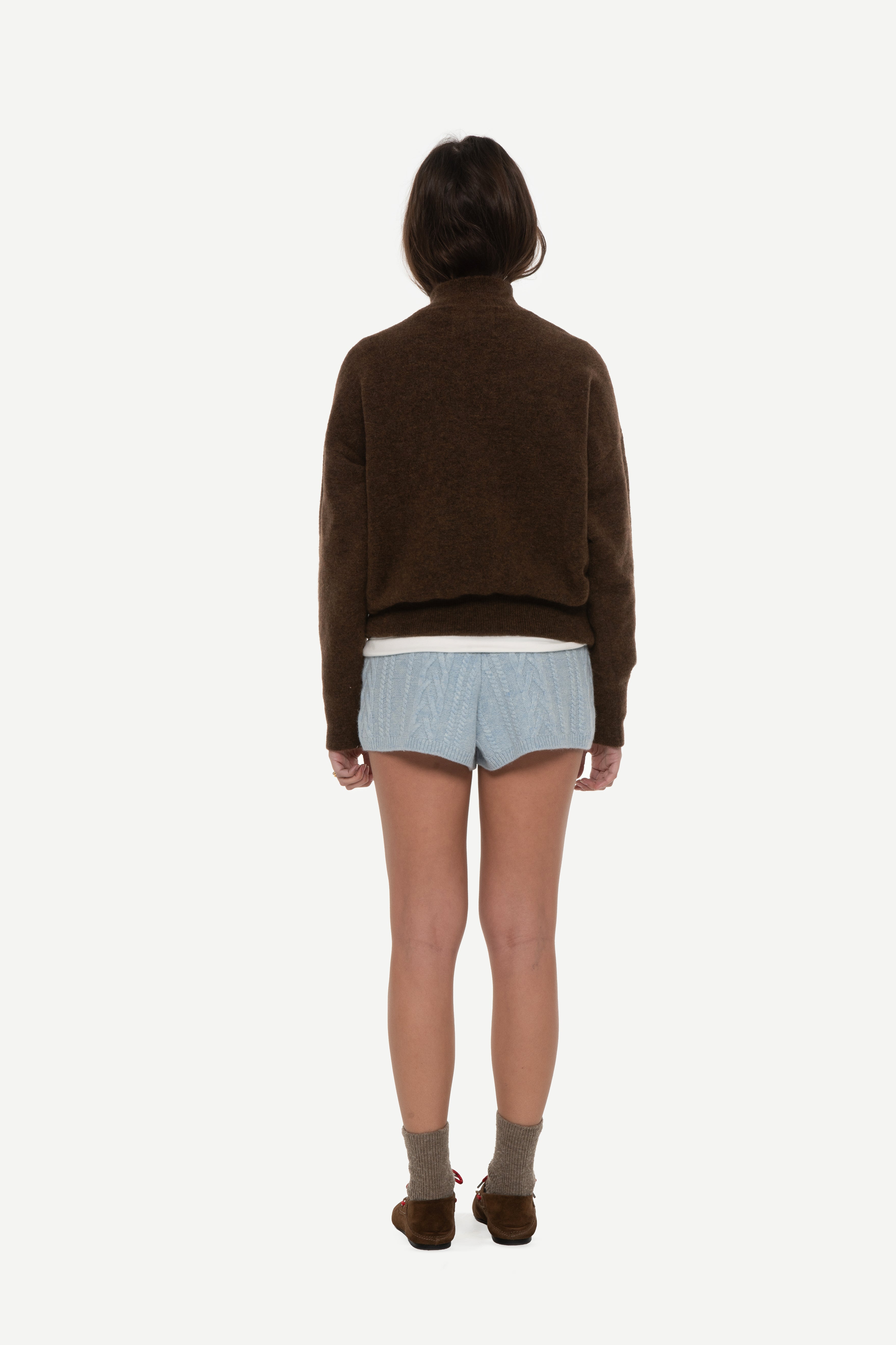 Back view of woman wearing a brown mockneck sweater and blue shorts on a white background