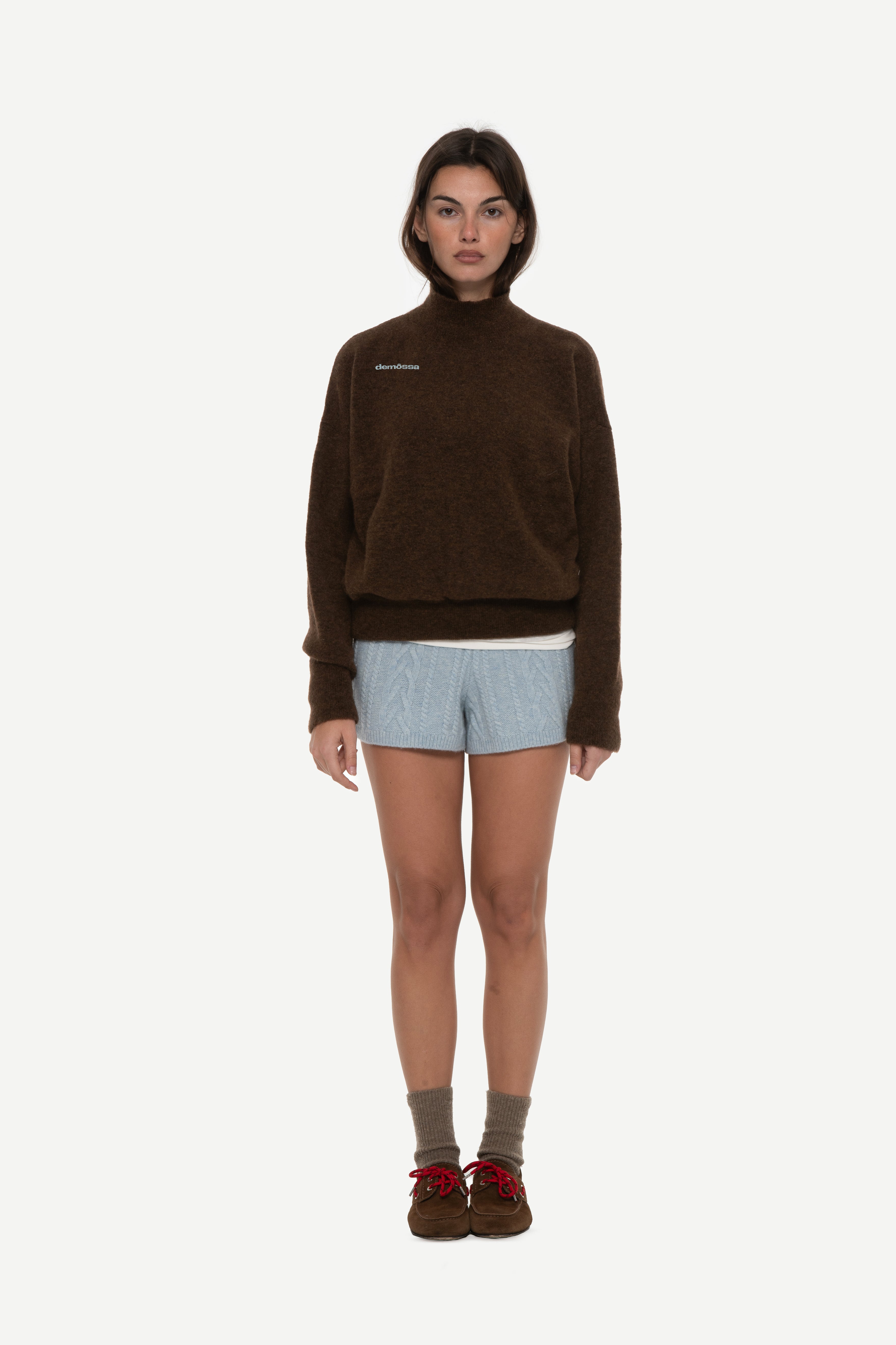 front view of woman wearing a brown mockneck sweater with demossa embroidered on upper chest and blue shorts on a white background