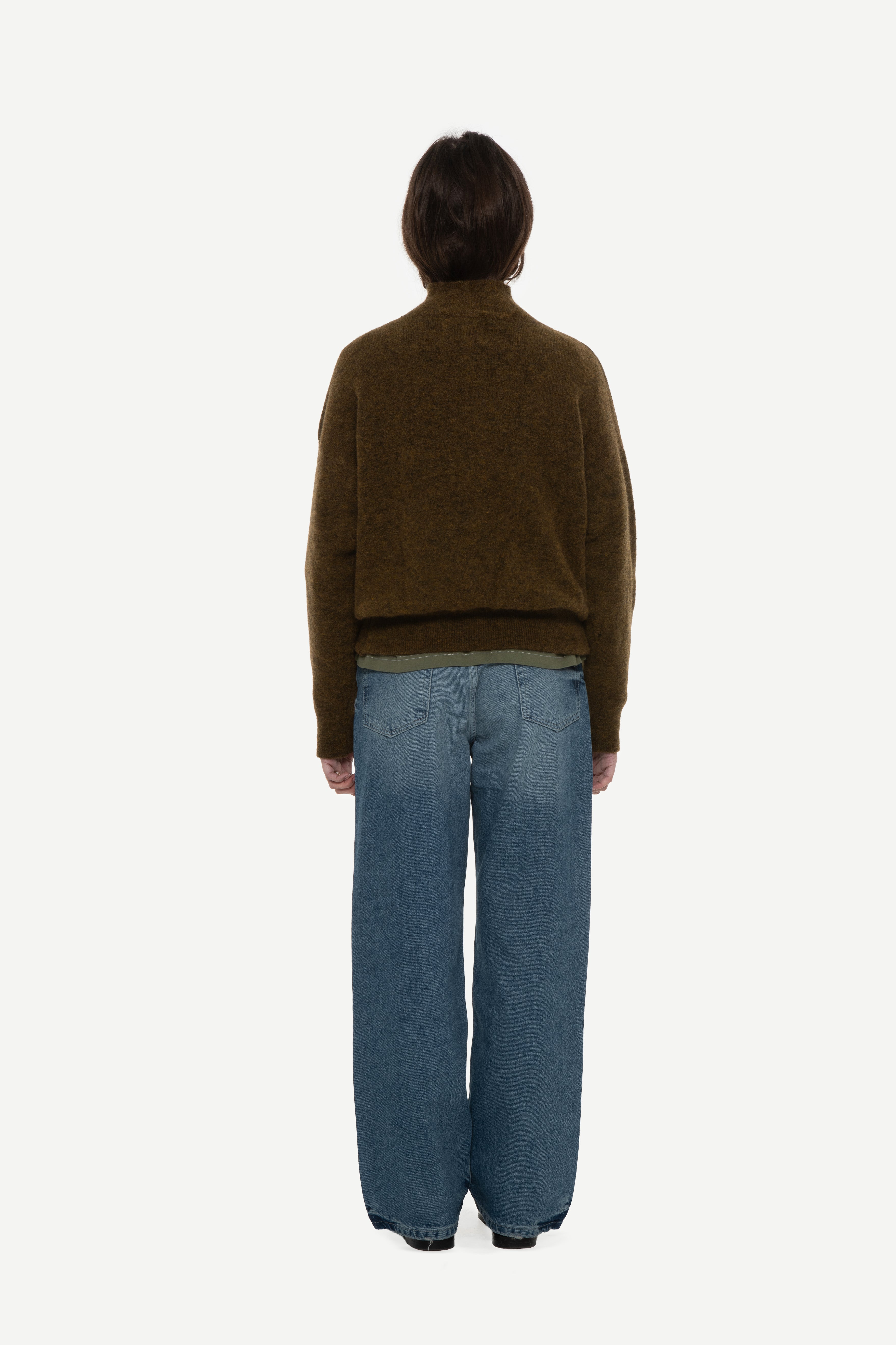 back view of woman wearing a olive mockneck sweater and blue jeans on a white background