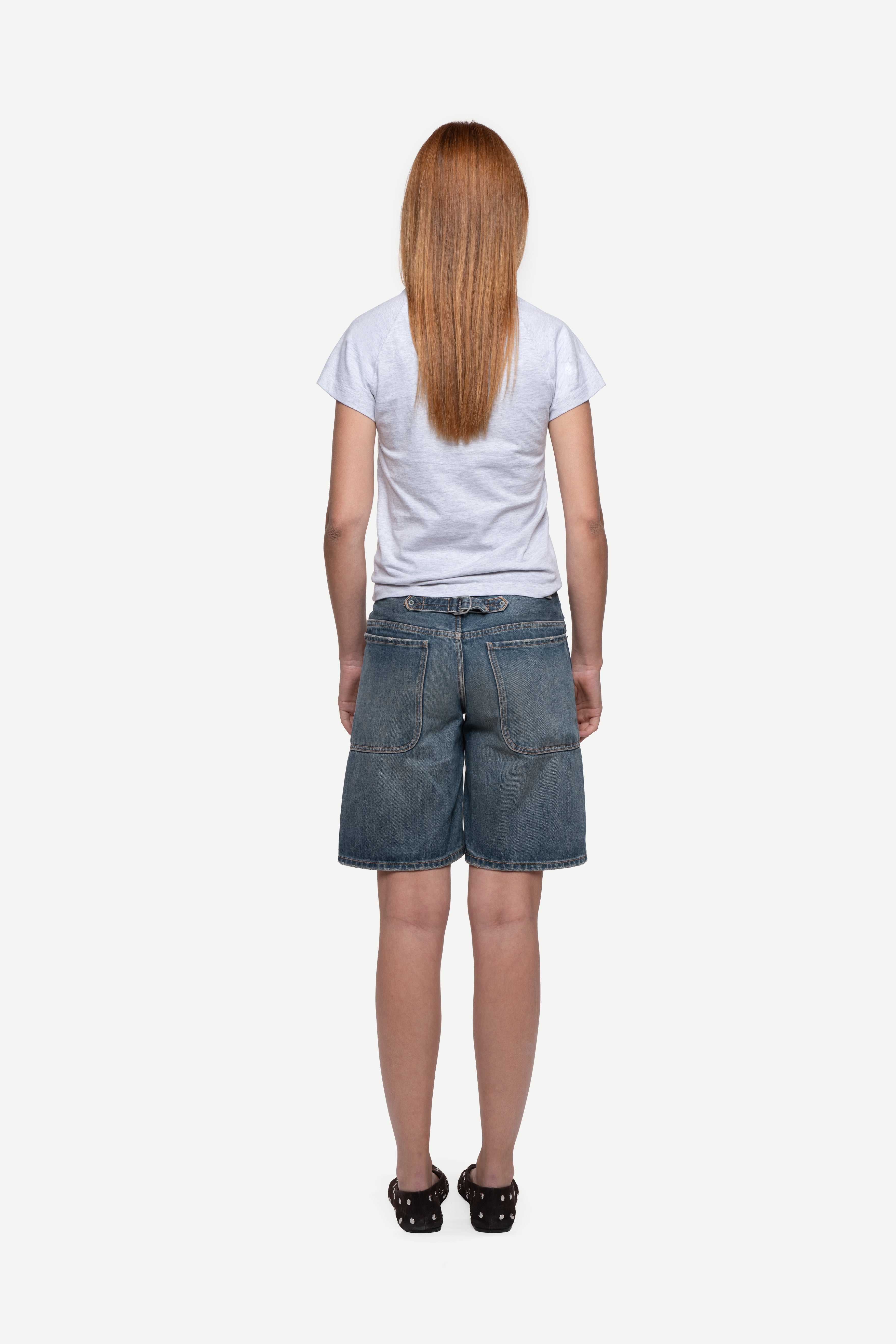back view of woman wearing a fitted short-sleeved t-shirt, blue short jeans and ballet flats on a white background