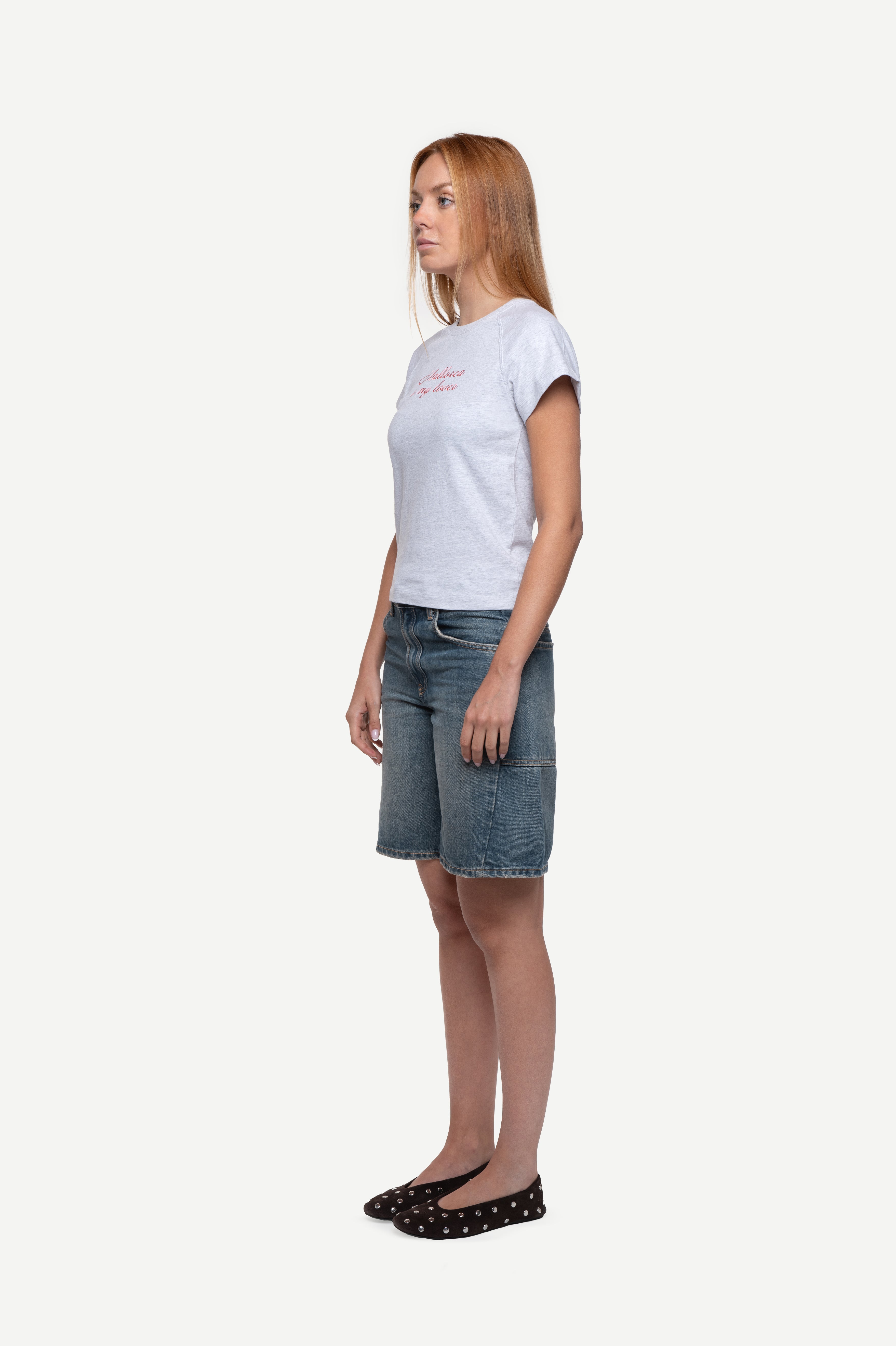 side view of woman wearing a fitted short-sleeved t-shirt with "Mallorca is my lover" front print in bright red, blue short jeans and ballet flats on a white background