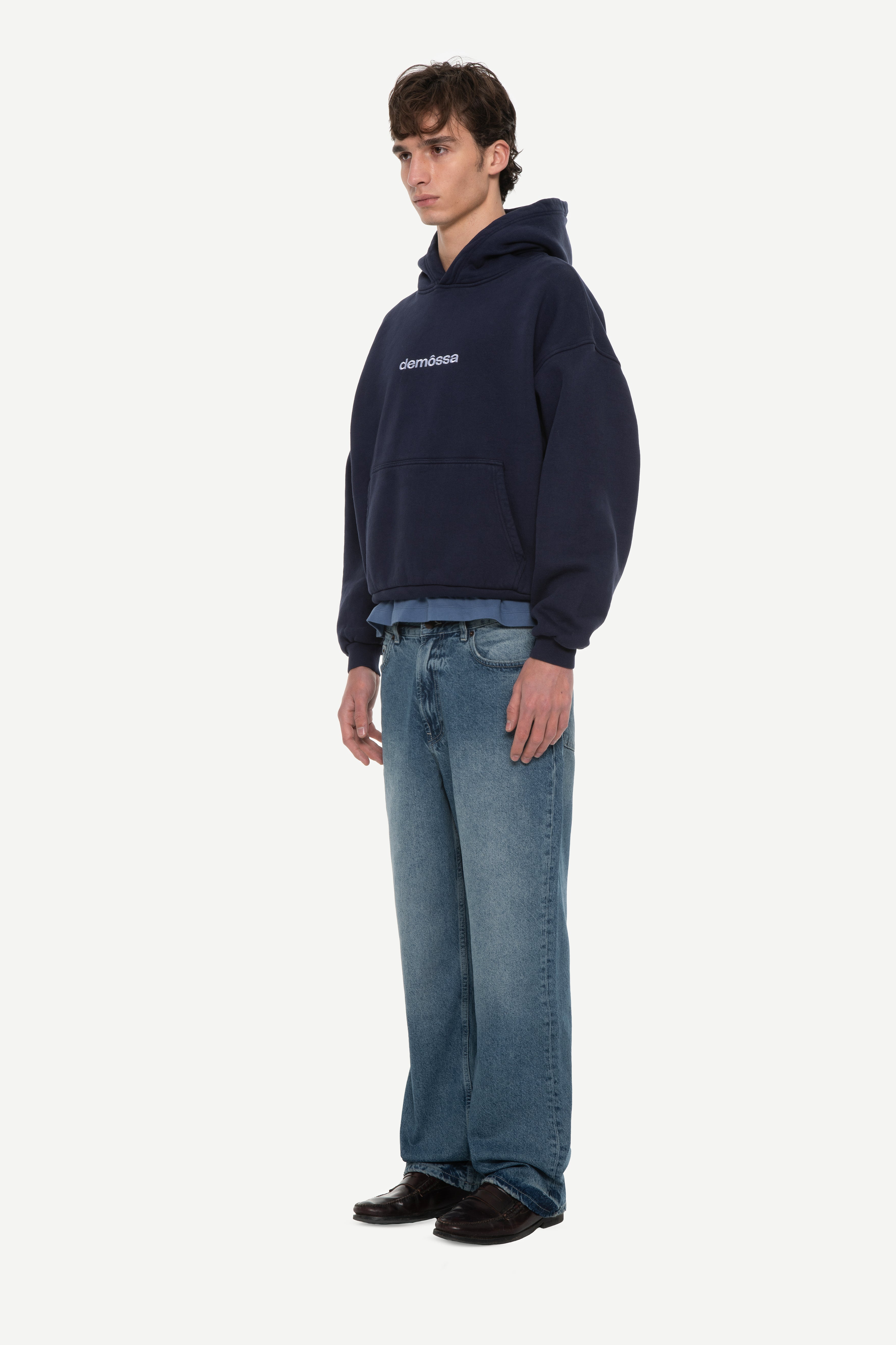 side view of man wearing a navy hoodie with “demôssa” logo embroidered on the middle chest and blue jeans on a white background