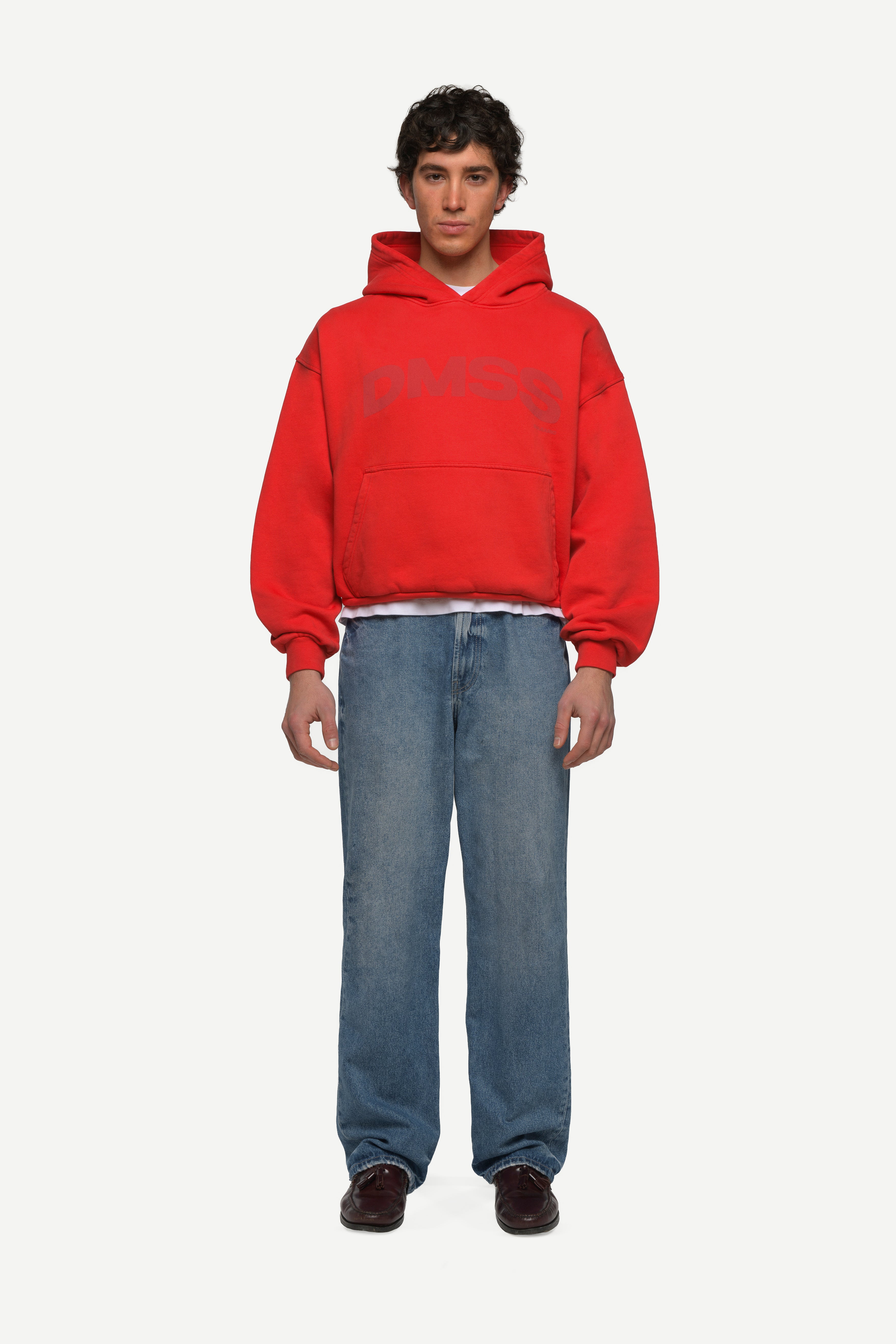 front view of man wearing a red hoodie with large DMSS logo in a subtle tone and blue jeans on a white background