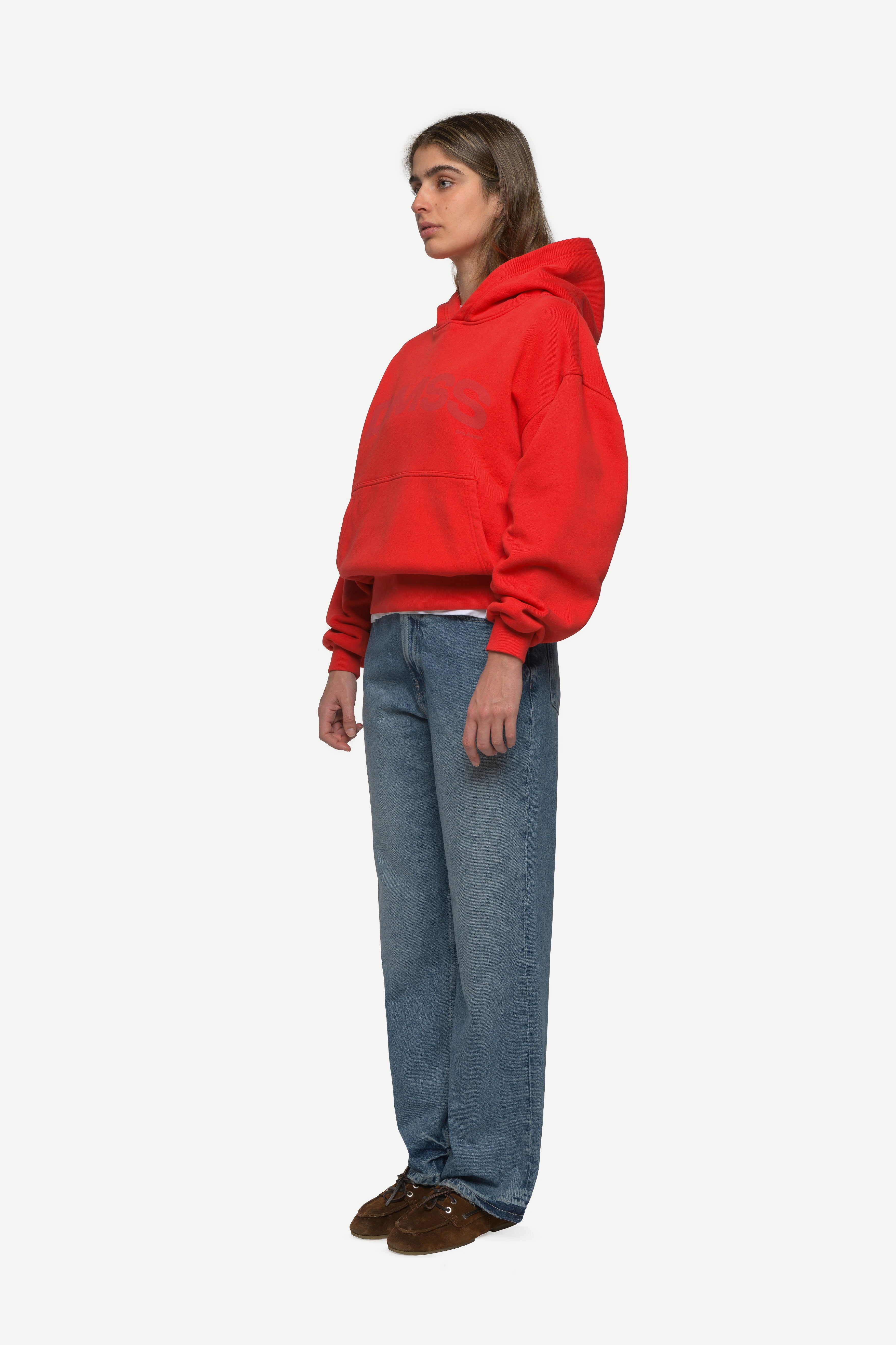 side view of woman wearing a red hoodie with large DMSS logo in a subtle tone and blue jeans on a white background