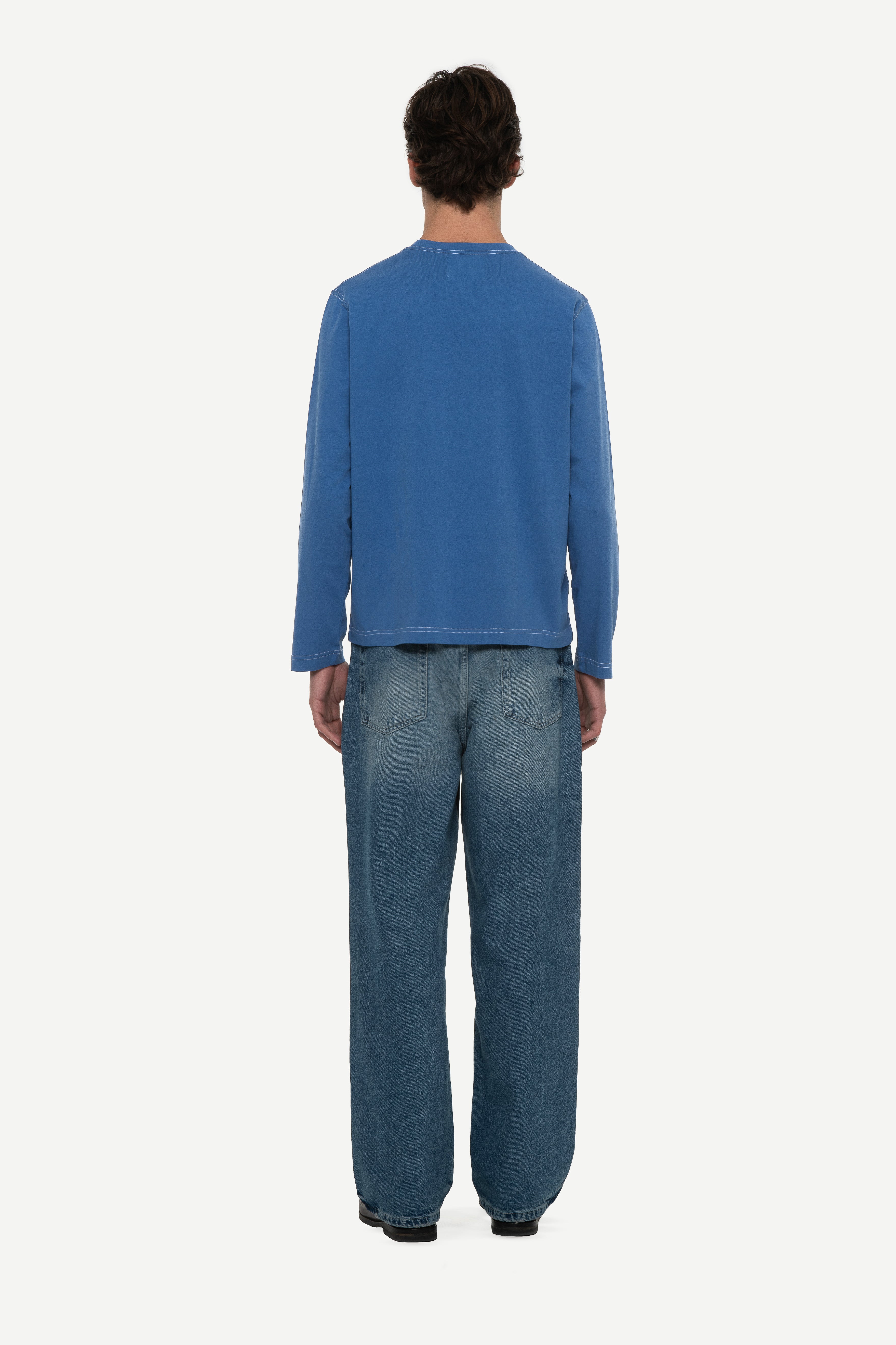 back view of man wearing a blue long sleeve tee with contrast stitching and blue jeans on a white background