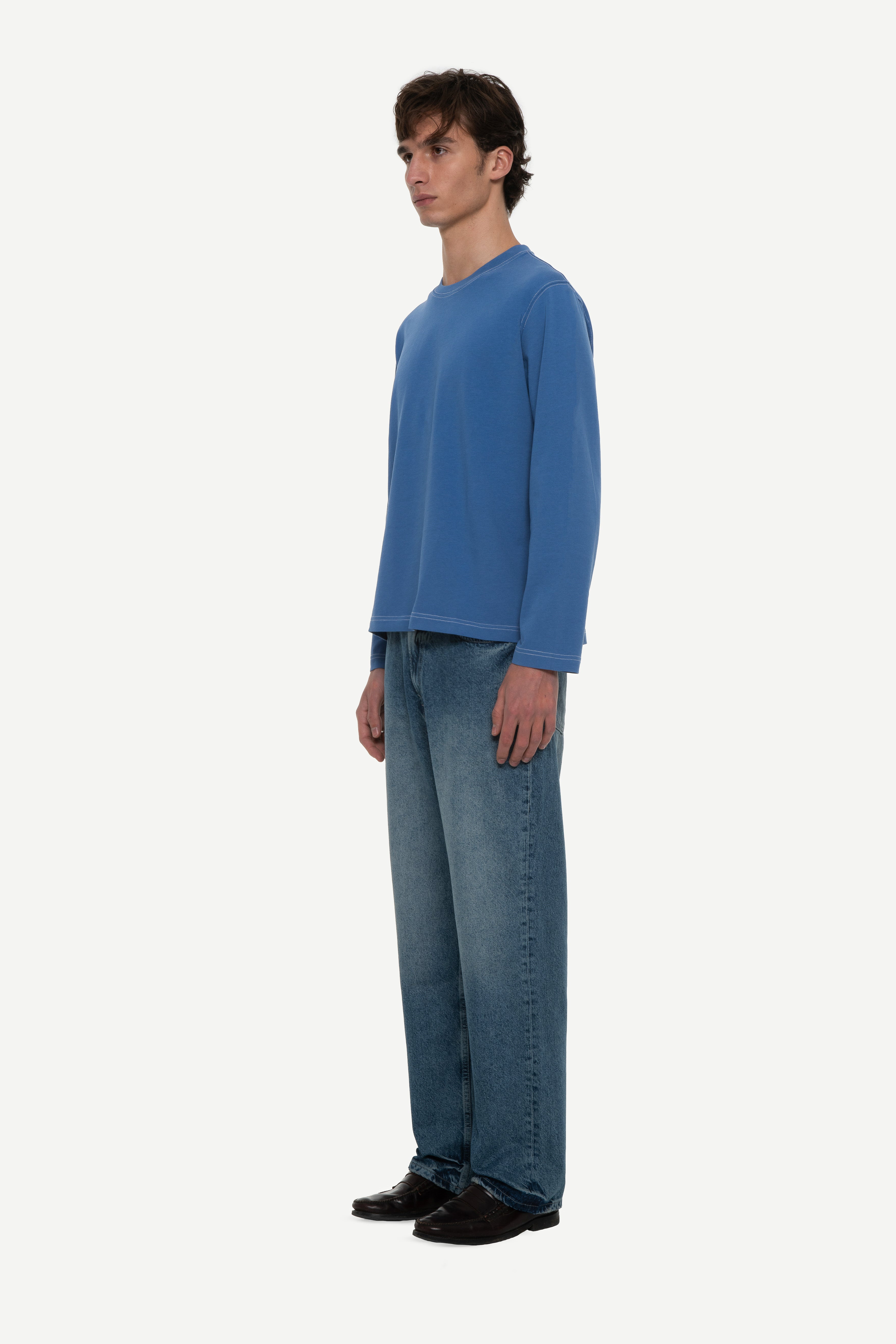 side view of man wearing a blue long sleeve tee with contrast stitching and blue jeans on a white background