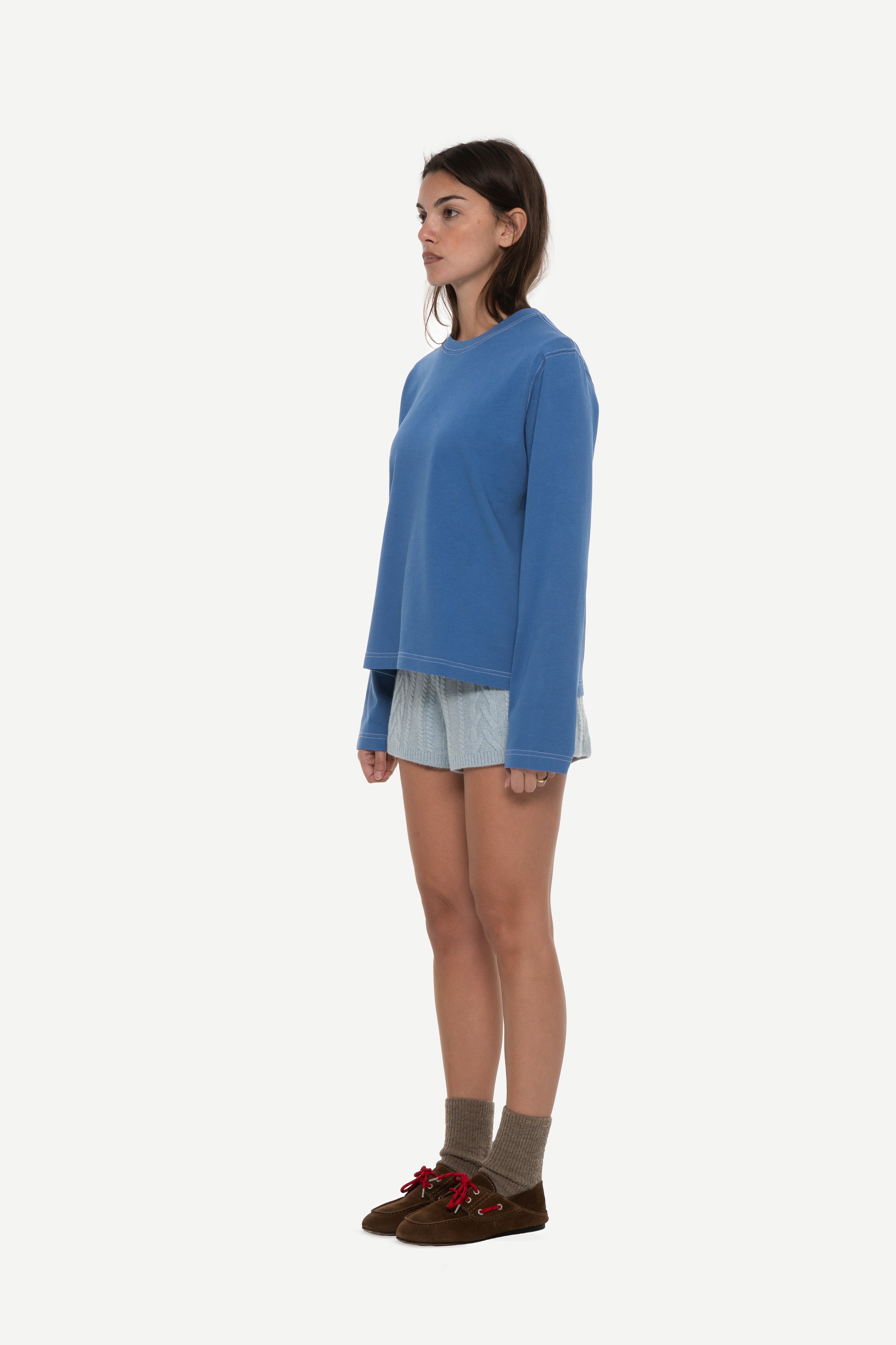 side view of woman wearing a blue long sleeve tee with contrast stitching and blue shorts on a white background