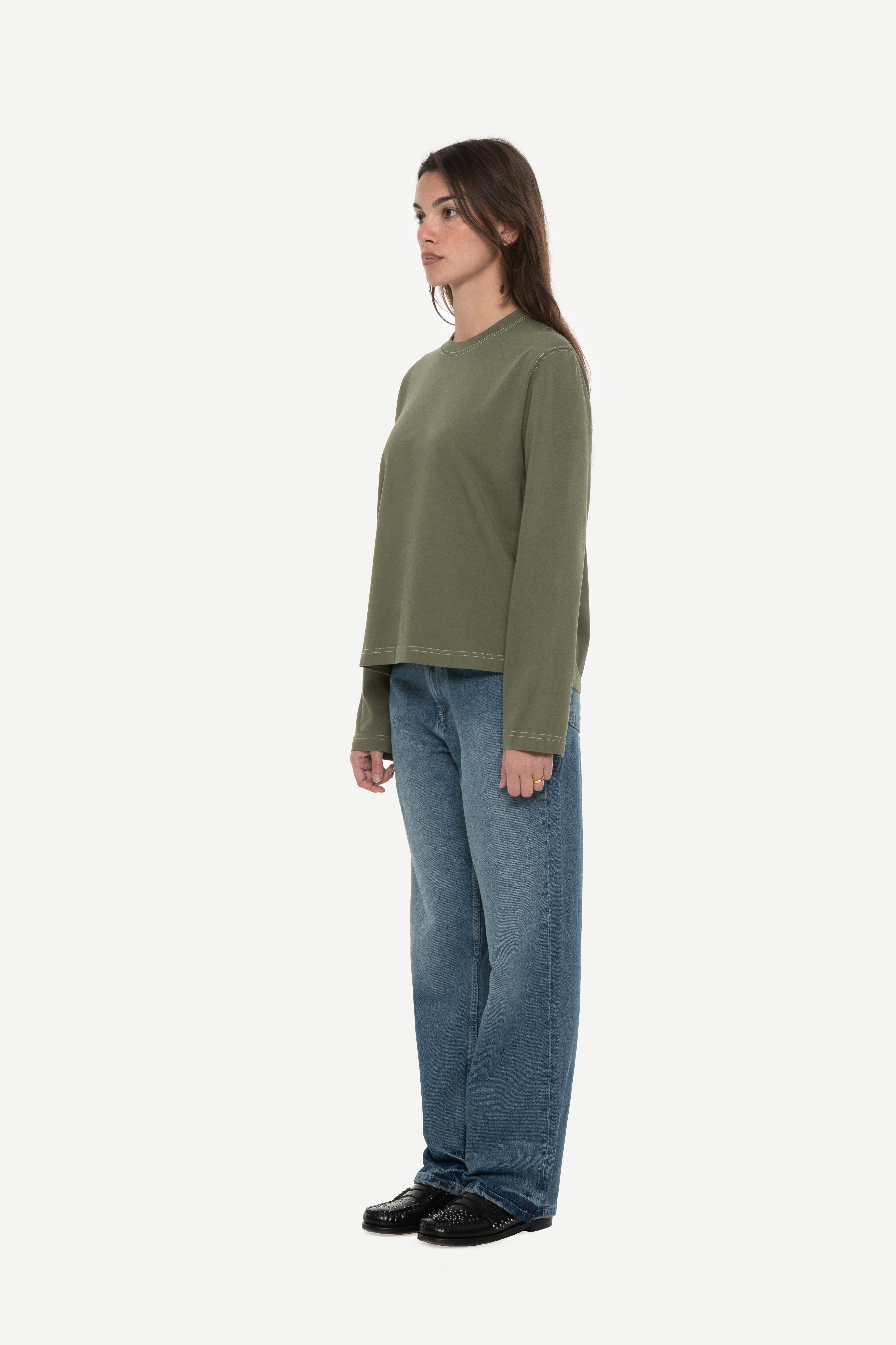 side view of woman wearing a military green long sleeve tee with contrast stitching and blue jeans on a white background