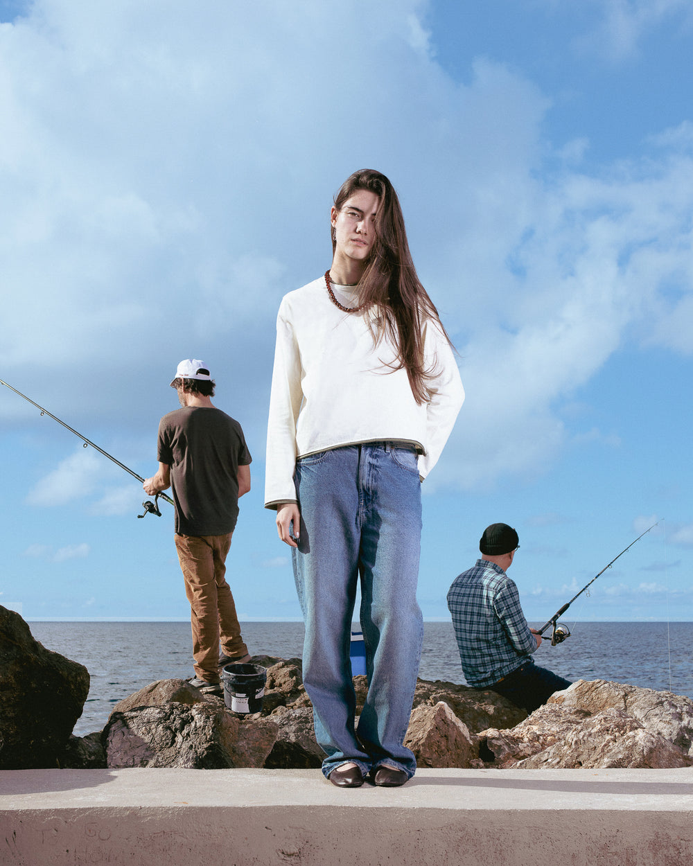 Person standing on rocks by the sea with two others fishing, under a blue sky.