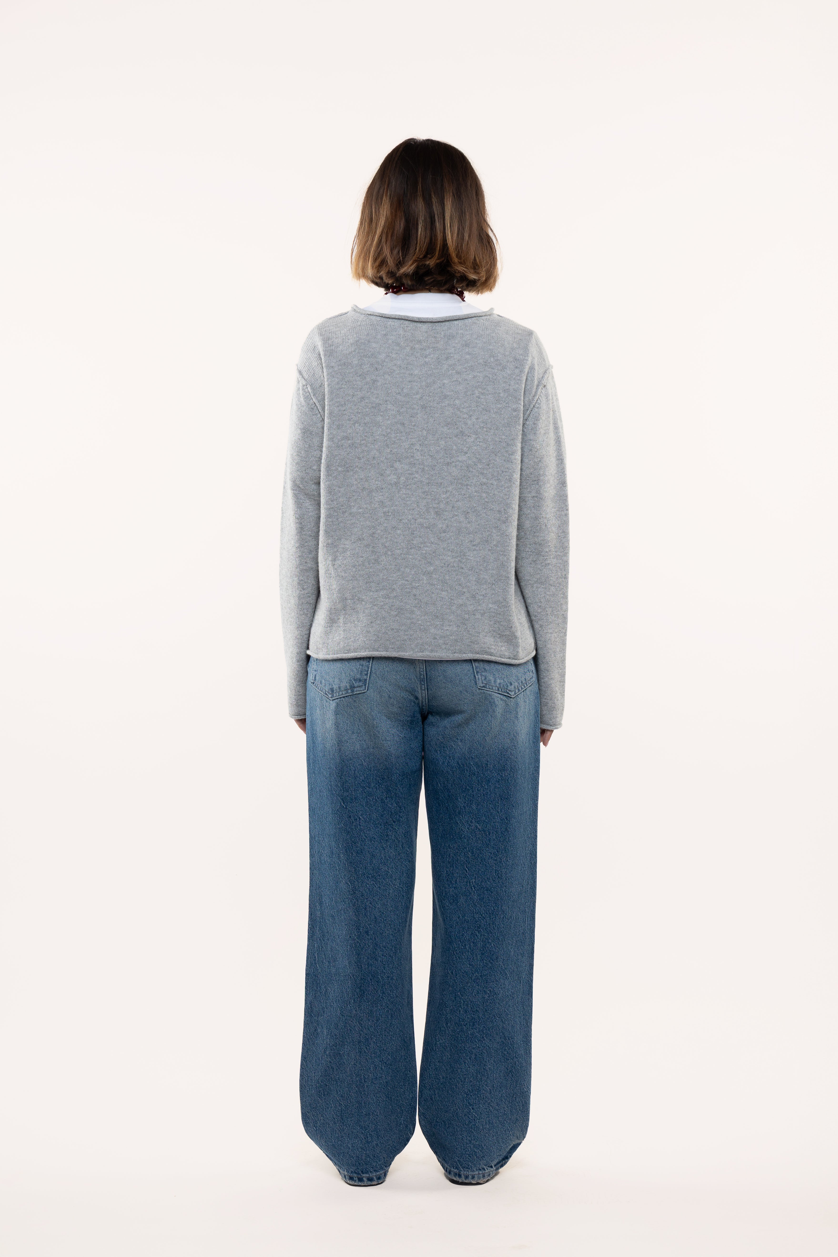 Back view of a woman wearing a light gray sweater and blue jeans on a white background