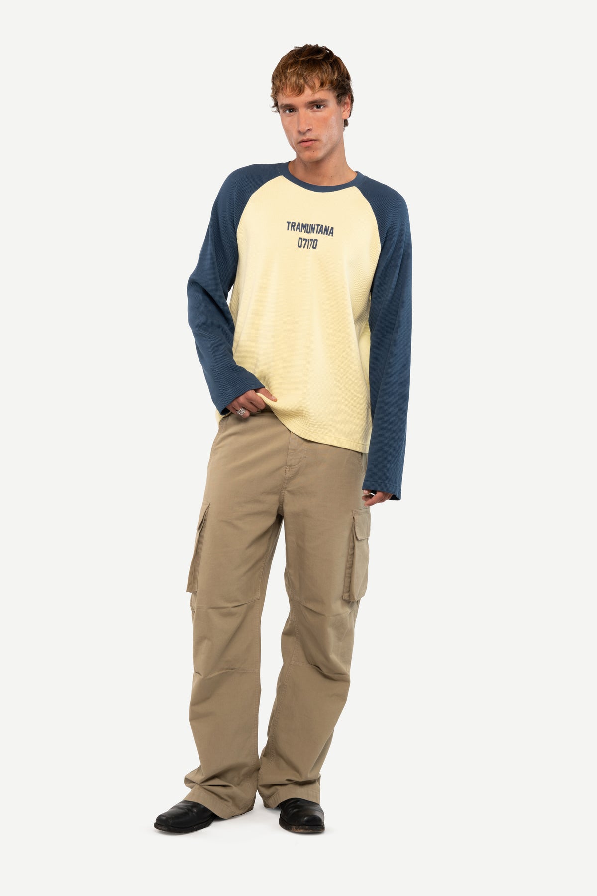 A male model wearing a cream and navy raglan long-sleeve shirt with 'TERRITORY 2070' printed on the chest, paired with beige cargo pants and black shoes, against a light gray background.