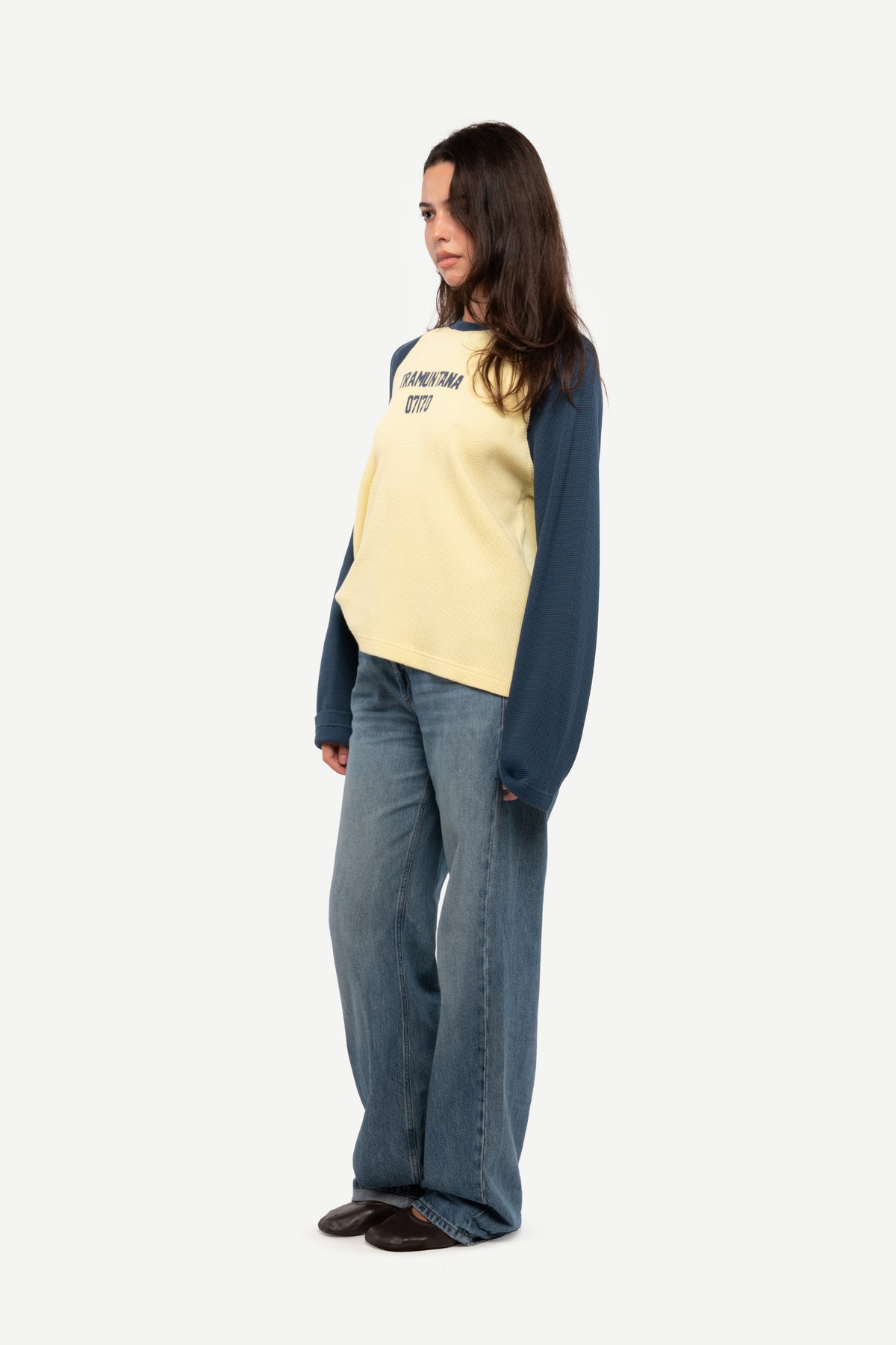 A woman modeling a two-tone raglan long-sleeve top with a pale yellow body and navy sleeves, paired with wide-leg blue jeans, shown against a plain light gray background.
