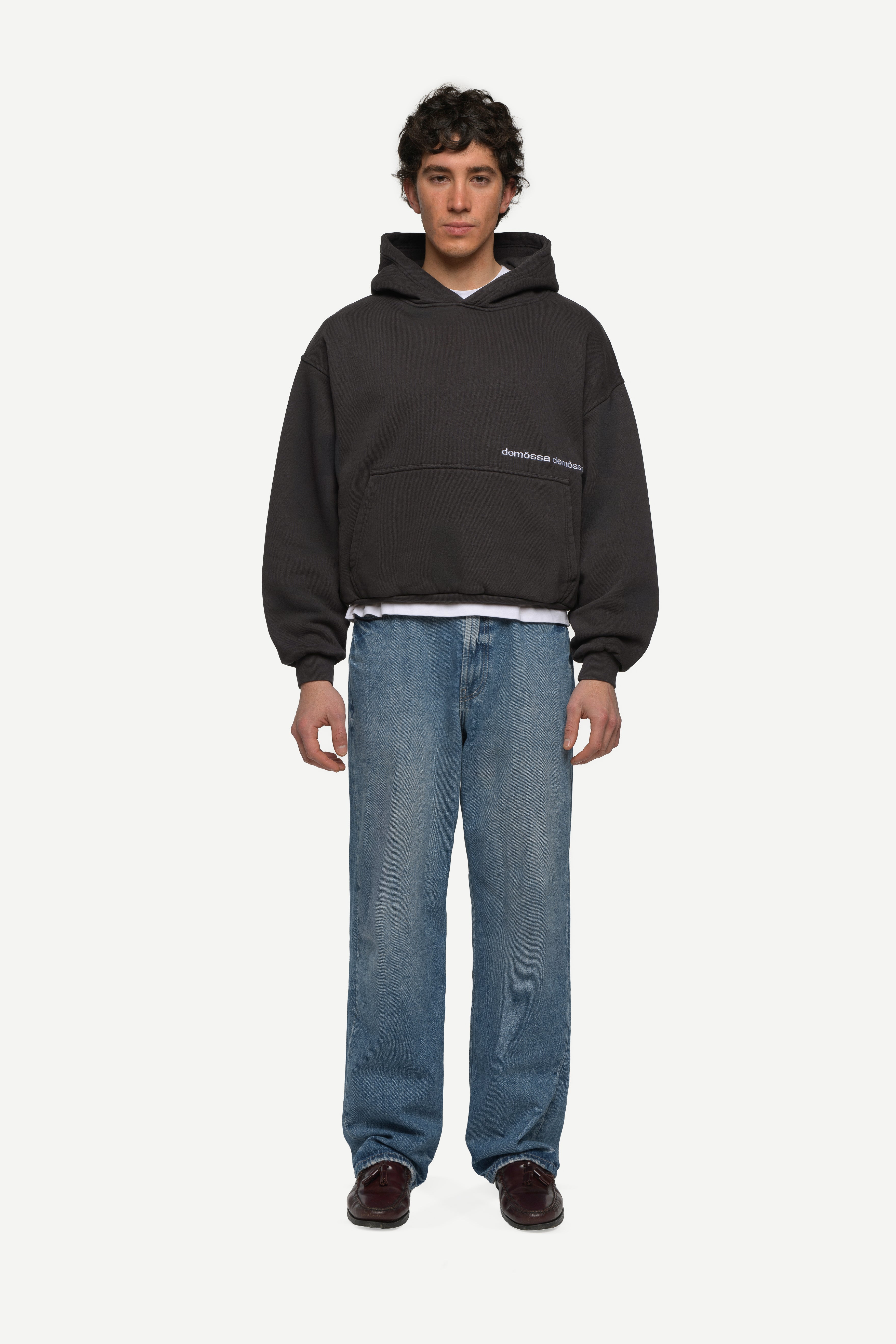 front view of man wearing a black hoodie with double demôssa logo on the side and blue jeans on a white background
