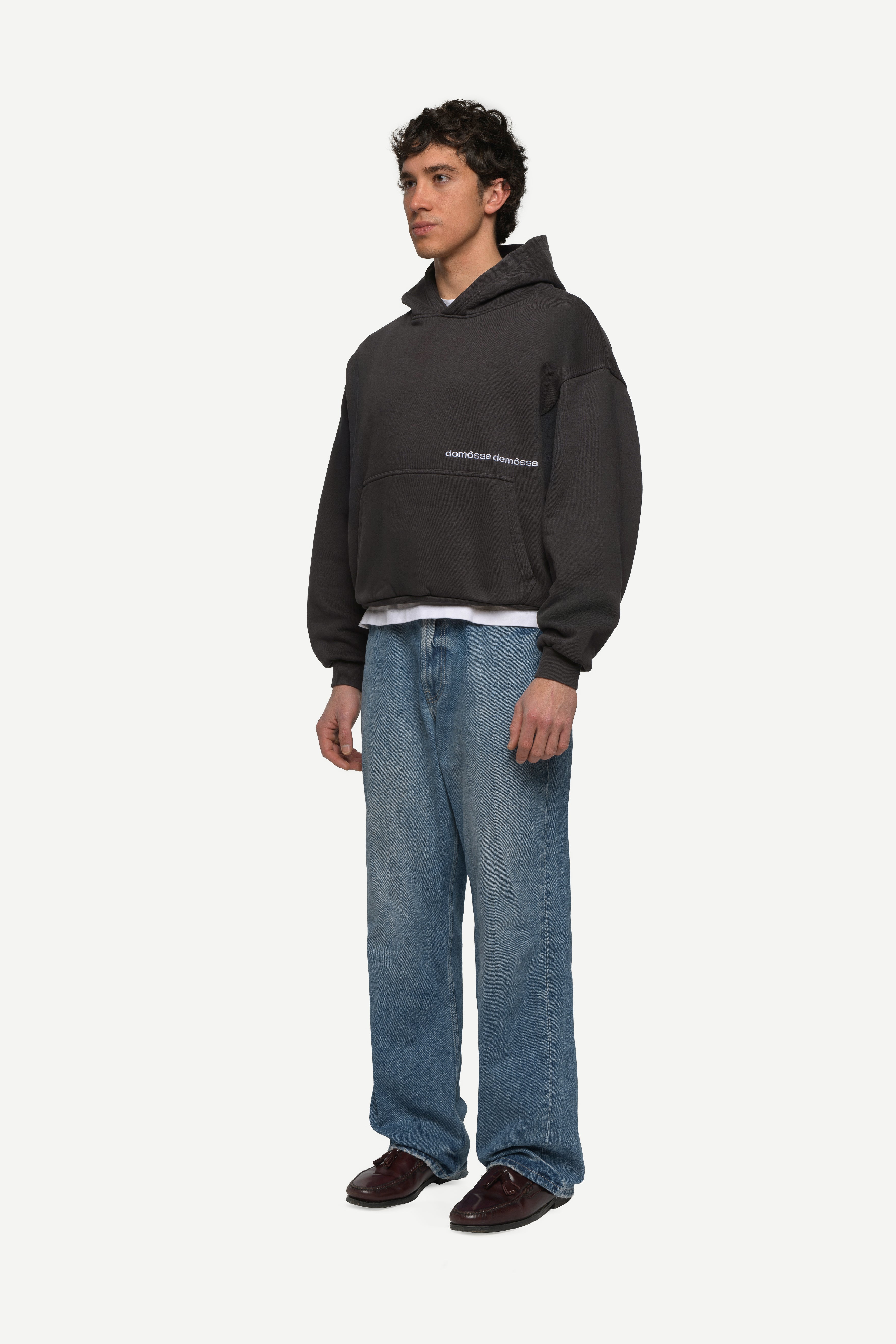 side view of man wearing a black hoodie with double demossa logo on the side and blue jeans on a white background