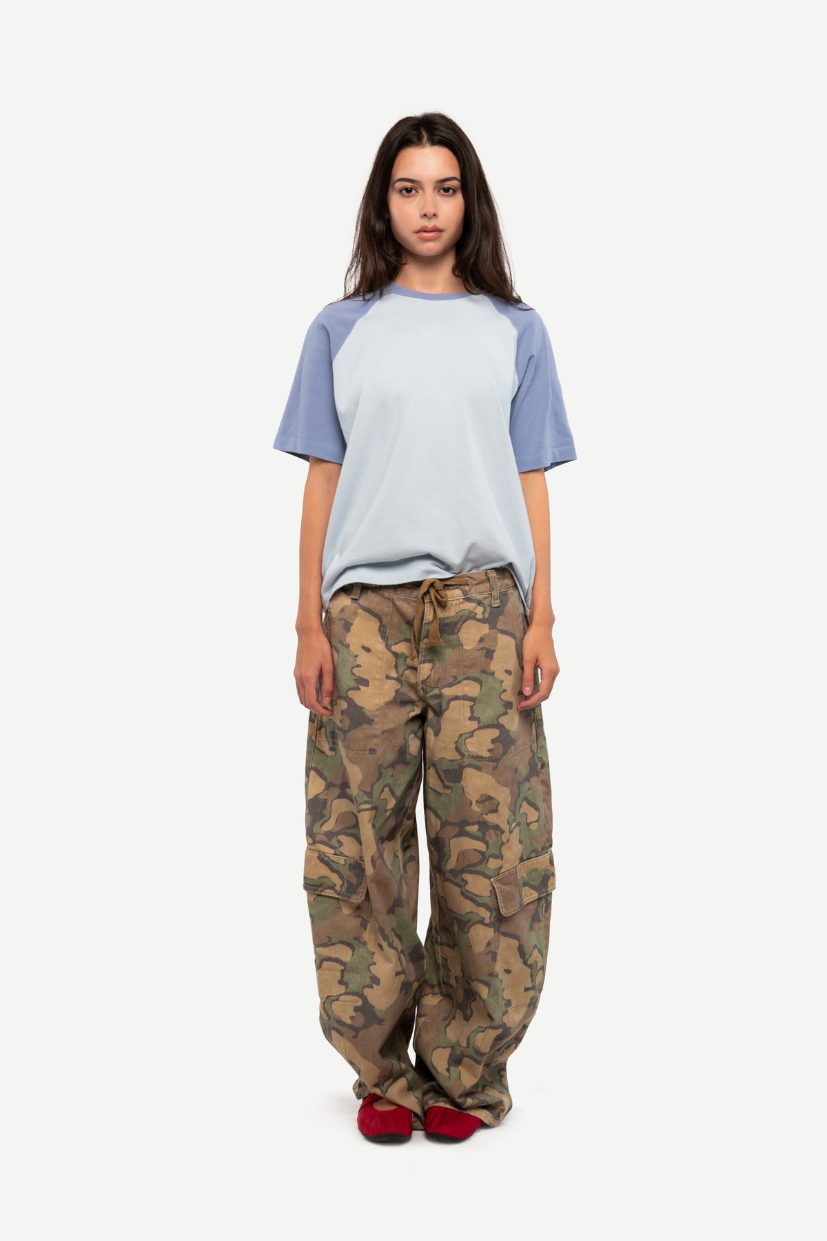 A model stands against a light gray background wearing a short-sleeve raglan t-shirt with a light blue body and muted blue sleeves, paired with loose camouflage cargo pants and red shoes.