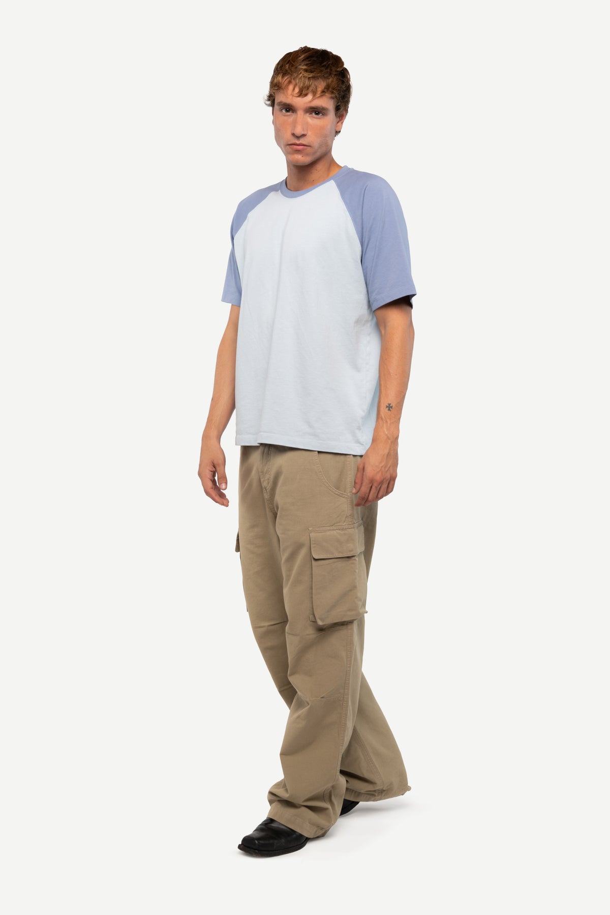 A man modeling a short-sleeve raglan t-shirt with a light blue body and lavender-blue sleeves, paired with beige cargo pants and black shoes, against a plain light gray background.