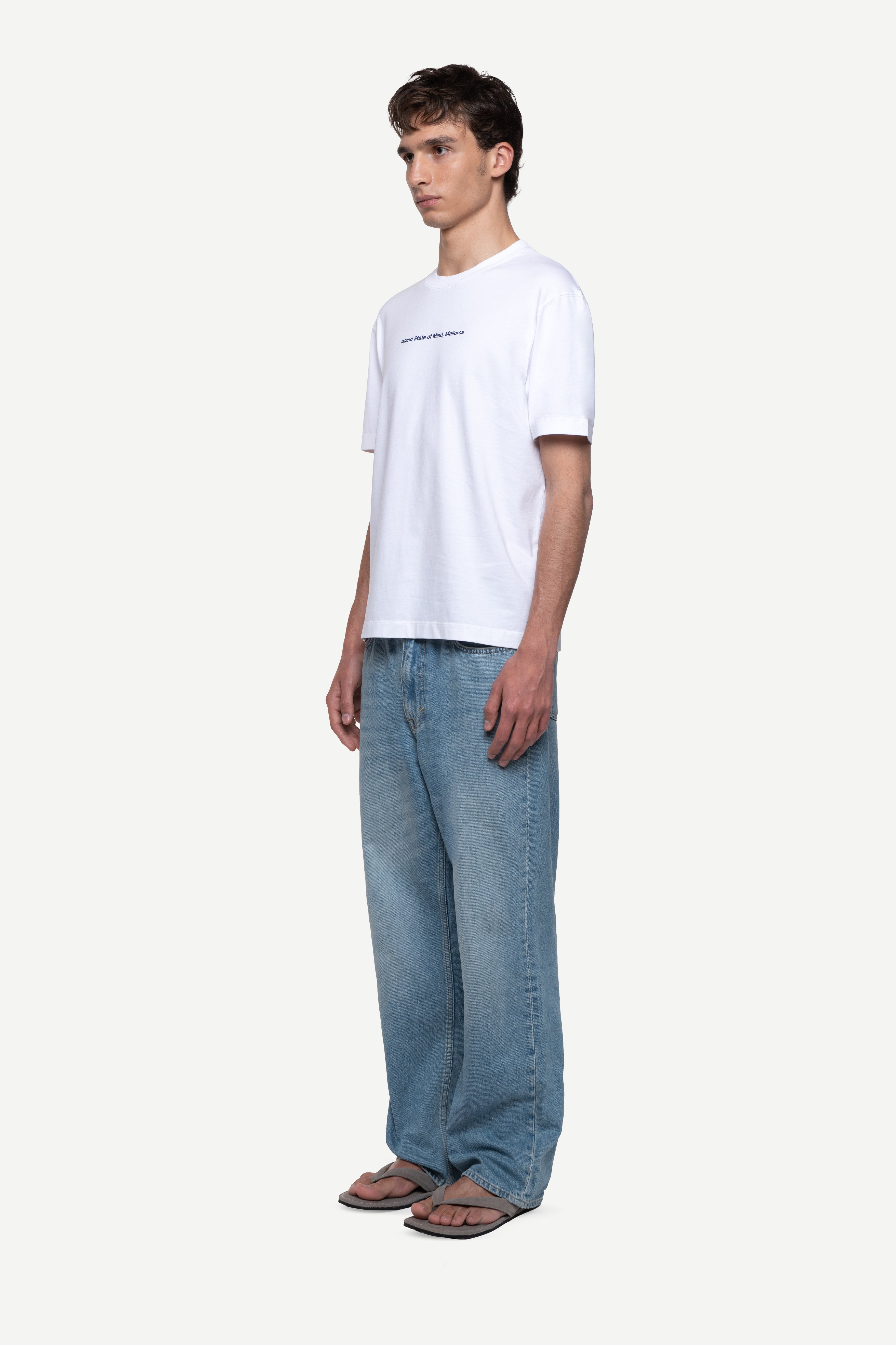 side view of man wearing a regular fit black short-sleeved t-shirt with white double-layered neckline, sleeves and hem, blue jeans and flip flops on a white background