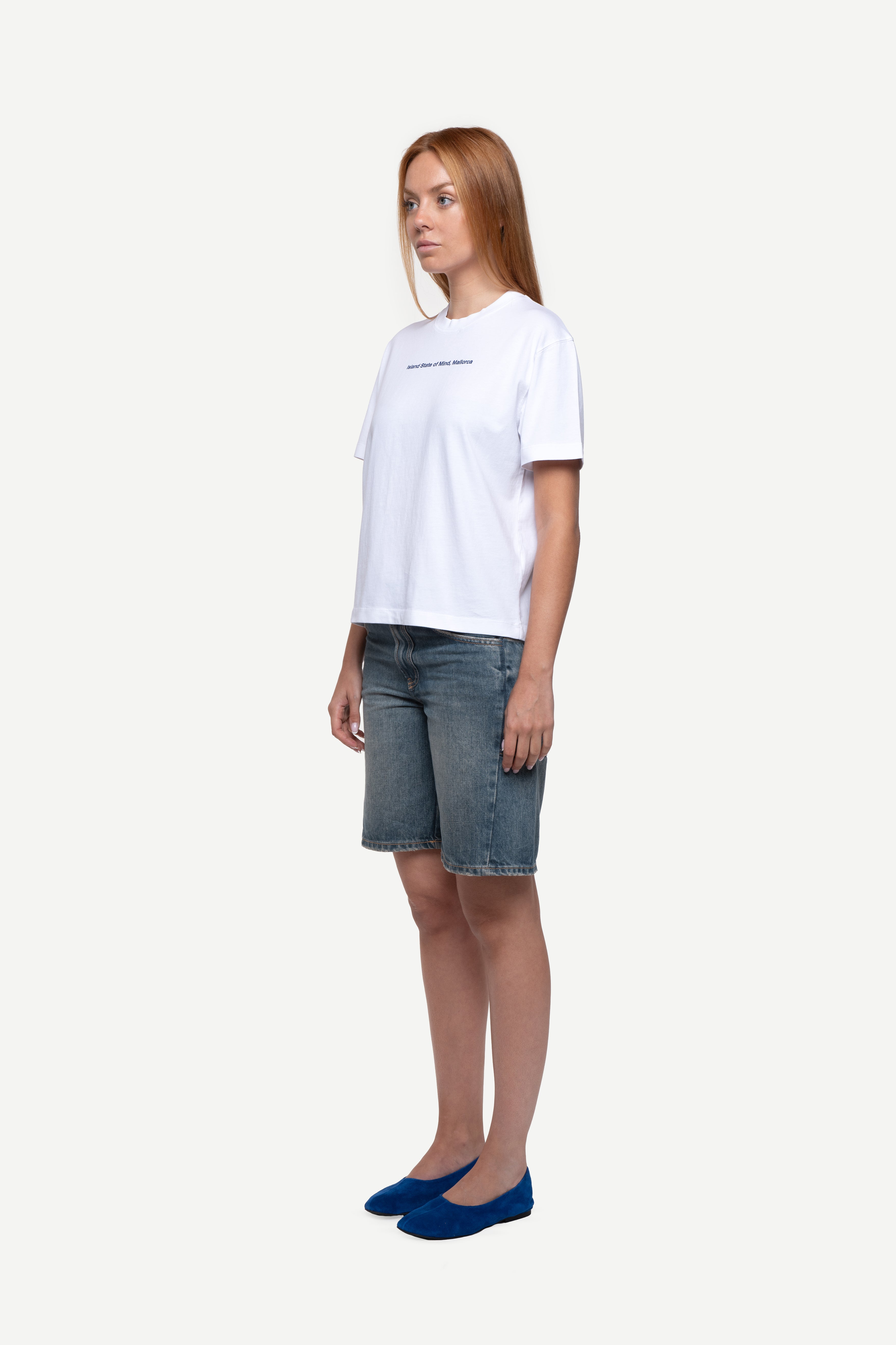 side view of woman wearing a regular fit black short-sleeved t-shirt with white double-layered neckline, sleeves and hem, blue jeans and flip flops on a white background