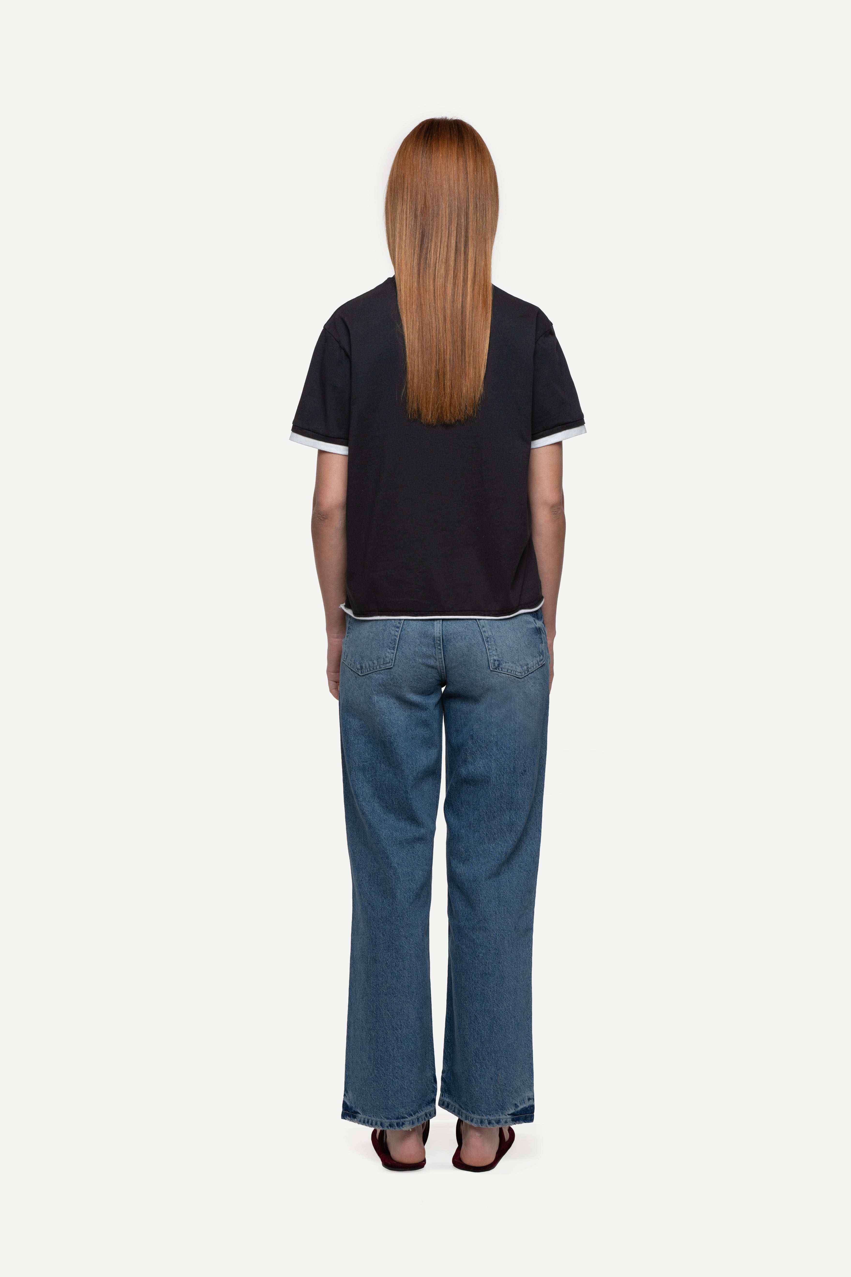 back view of woman wearing a regular fit black short-sleeved t-shirt, blue jeans and flip flops on a white background