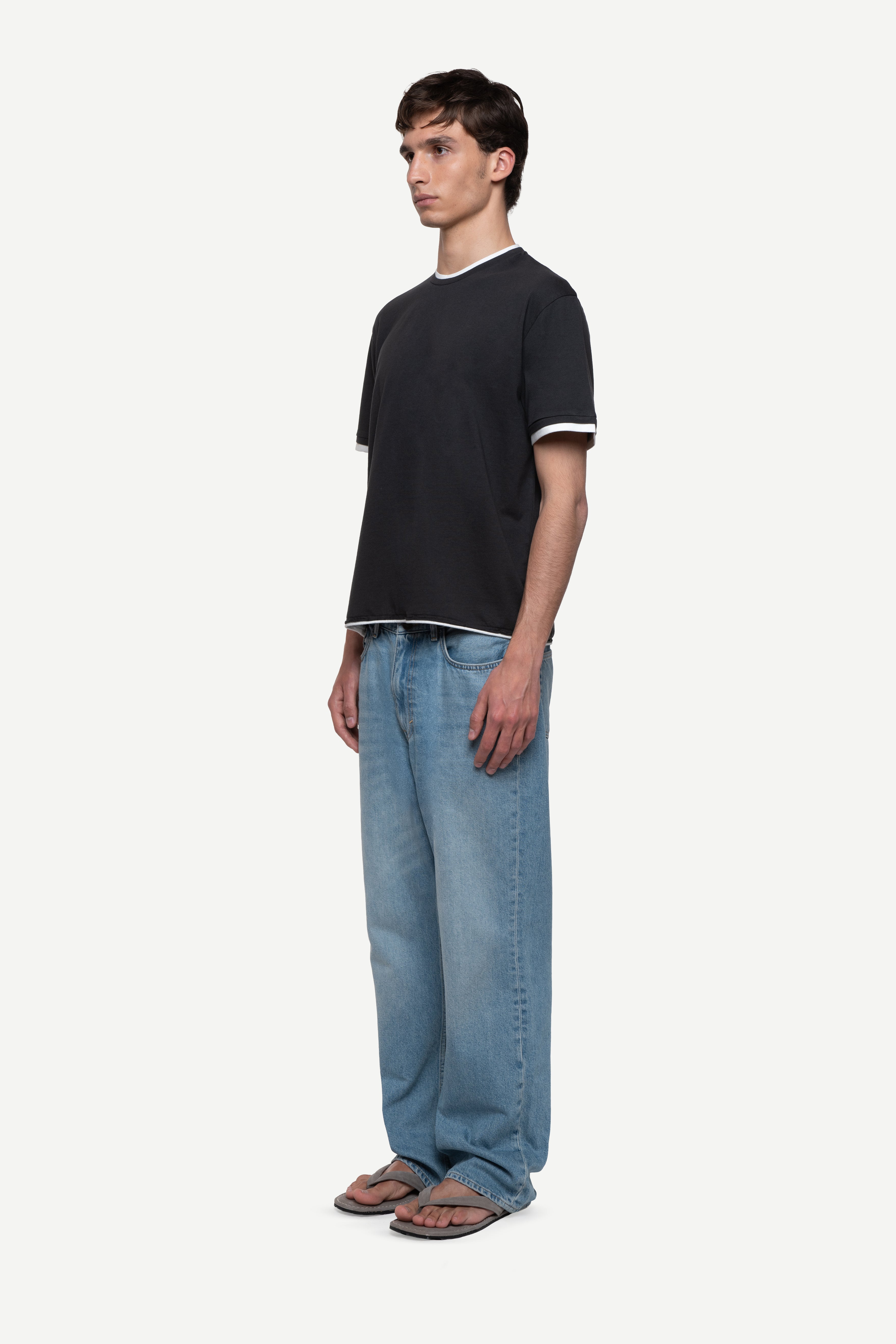 side view of man wearing a regular fit black short-sleeved t-shirt with white double-layered neckline, sleeves and hem, blue jeans and flip flops on a white background