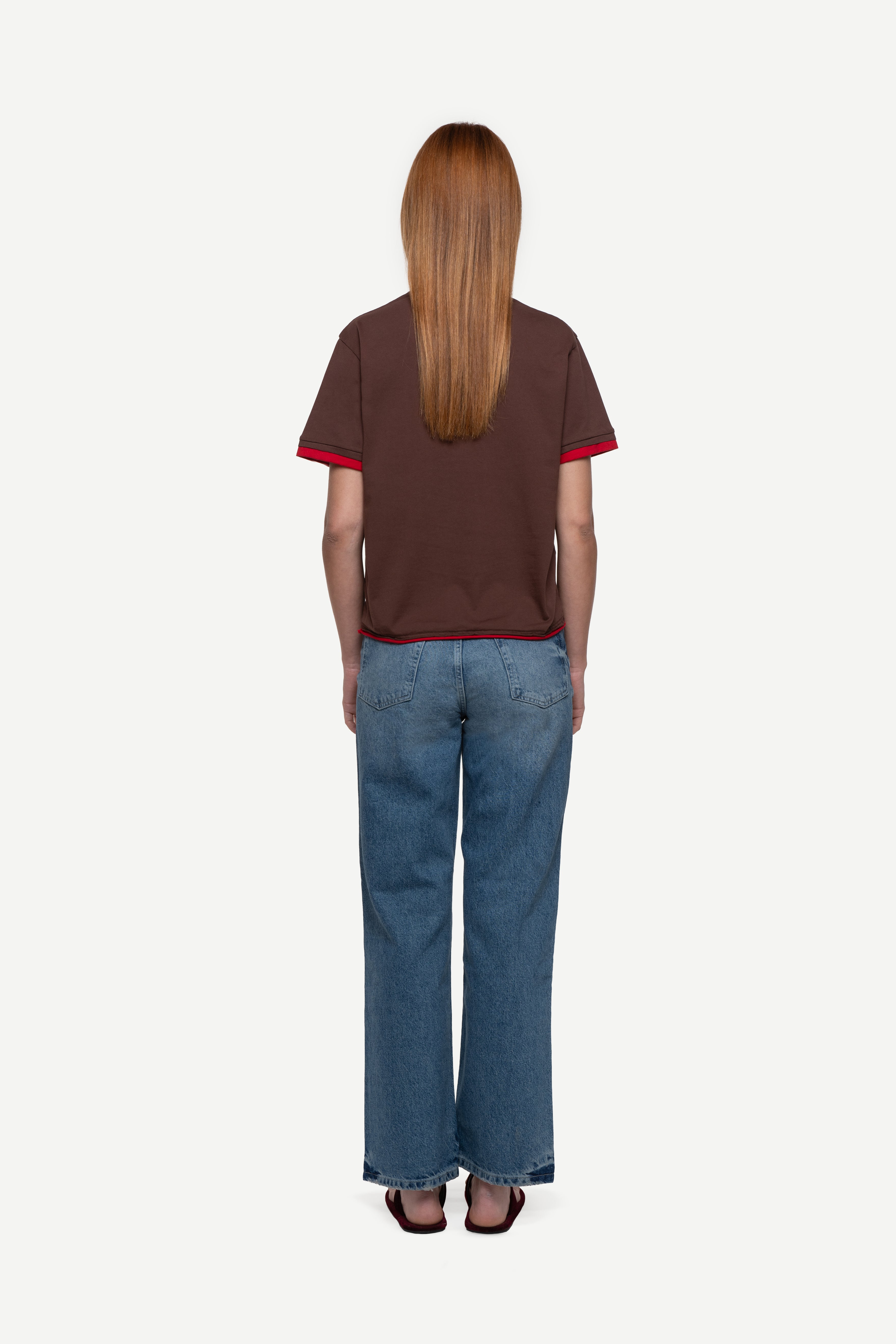 back view of woman wearing a regular fit brown short-sleeved t-shirt, blue jeans and flip flops on a white background