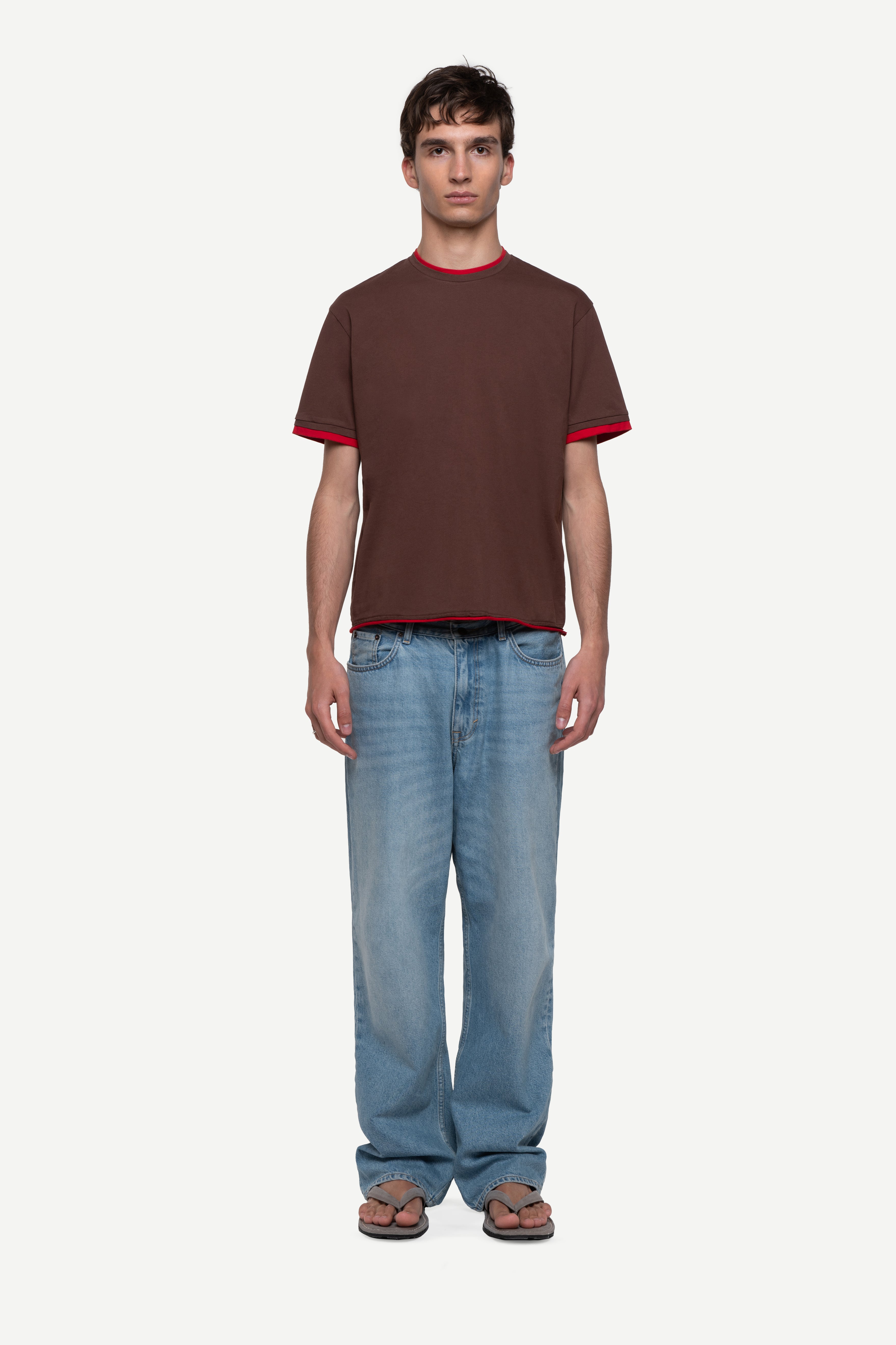 front view of man wearing a regular fit brown short-sleeved t-shirt with red double-layered neckline, sleeves and hem, blue jeans and flip flops on a white background