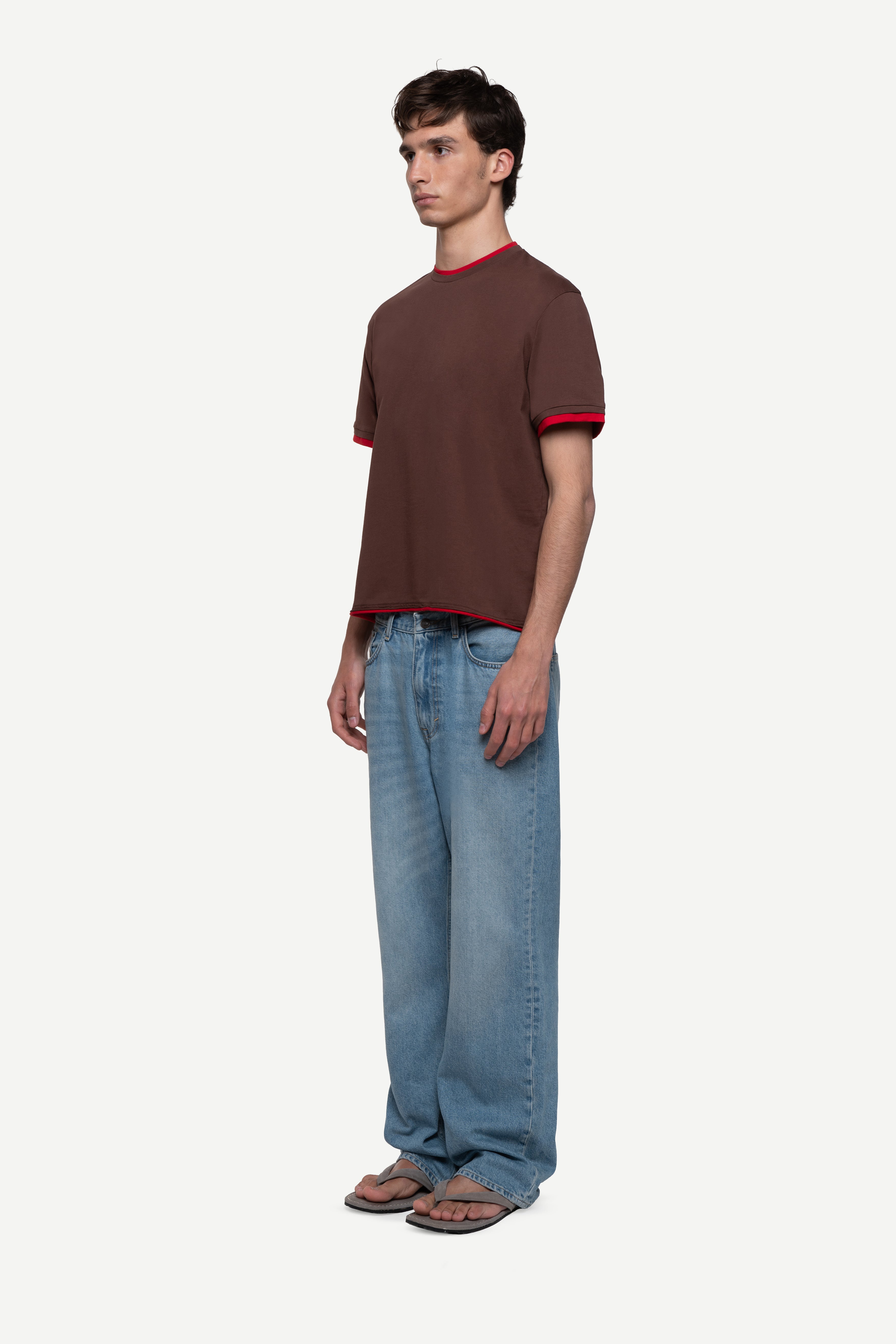 side view of man wearing a regular fit brown short-sleeved t-shirt with red double-layered neckline, sleeves and hem, blue jeans and flip flops on a white background