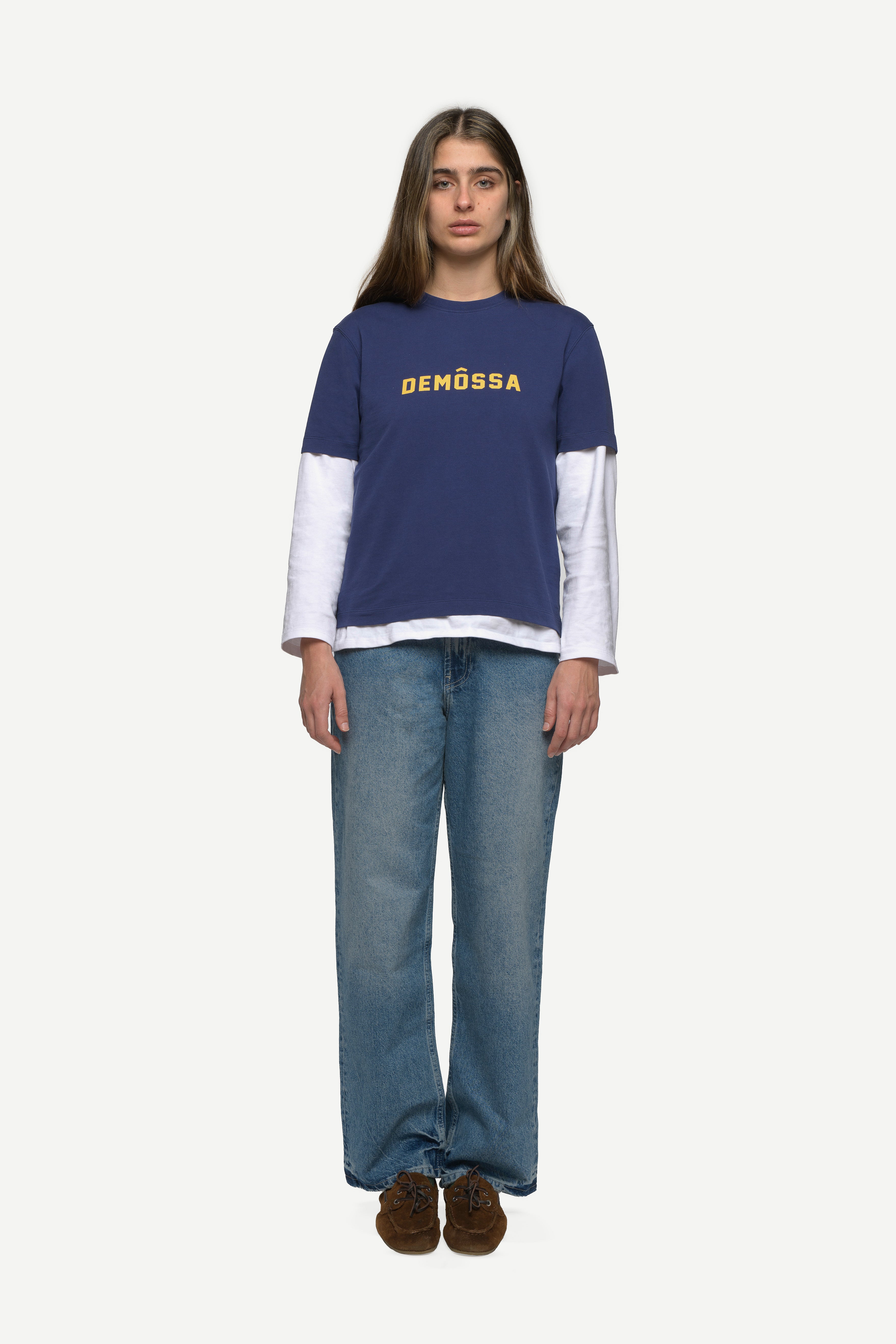 front view of woman wearing a navy t-shirt with yellow demossa logo on the chest and blue jeans on a white background